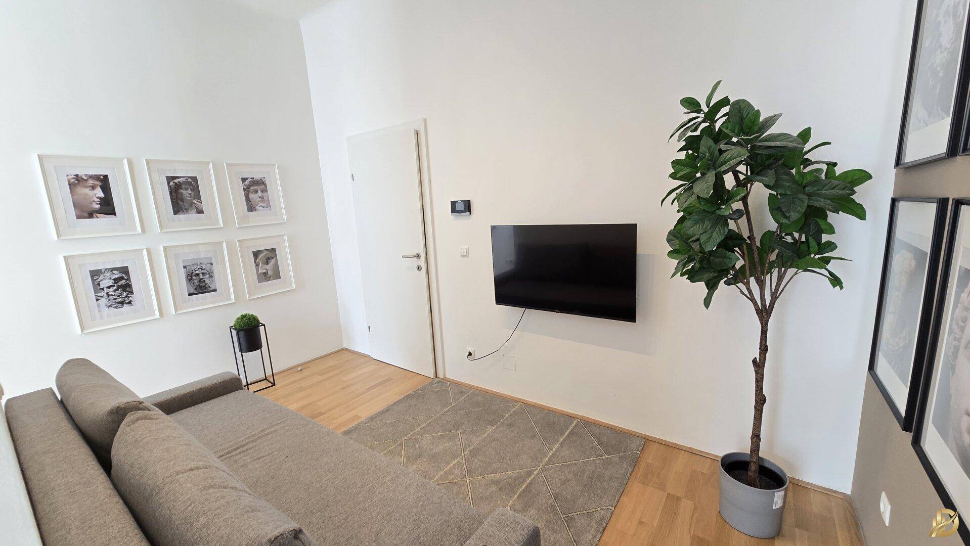 Wohnbereich mit Wandfernseher, beigem Sofa und dekorativer Pflanze, heller Holzboden.