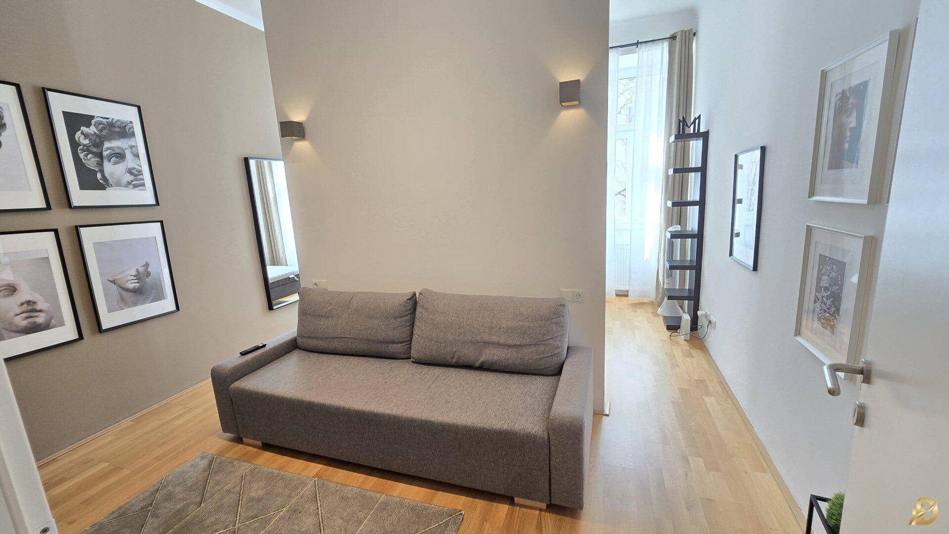 Wohnbereich mit beigem Sofa und gerahmten Bildern, offener Blick zum Schlafzimmer.