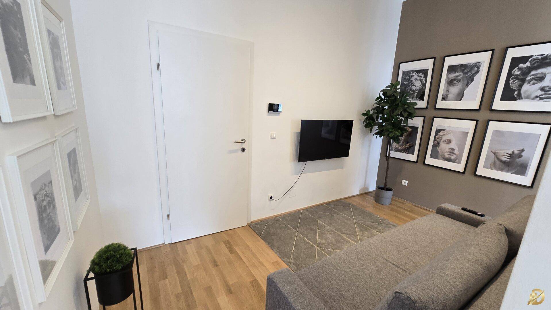 Wohnbereich mit Wandfernseher, beigem Sofa und Kunstwerken, heller Holzboden.