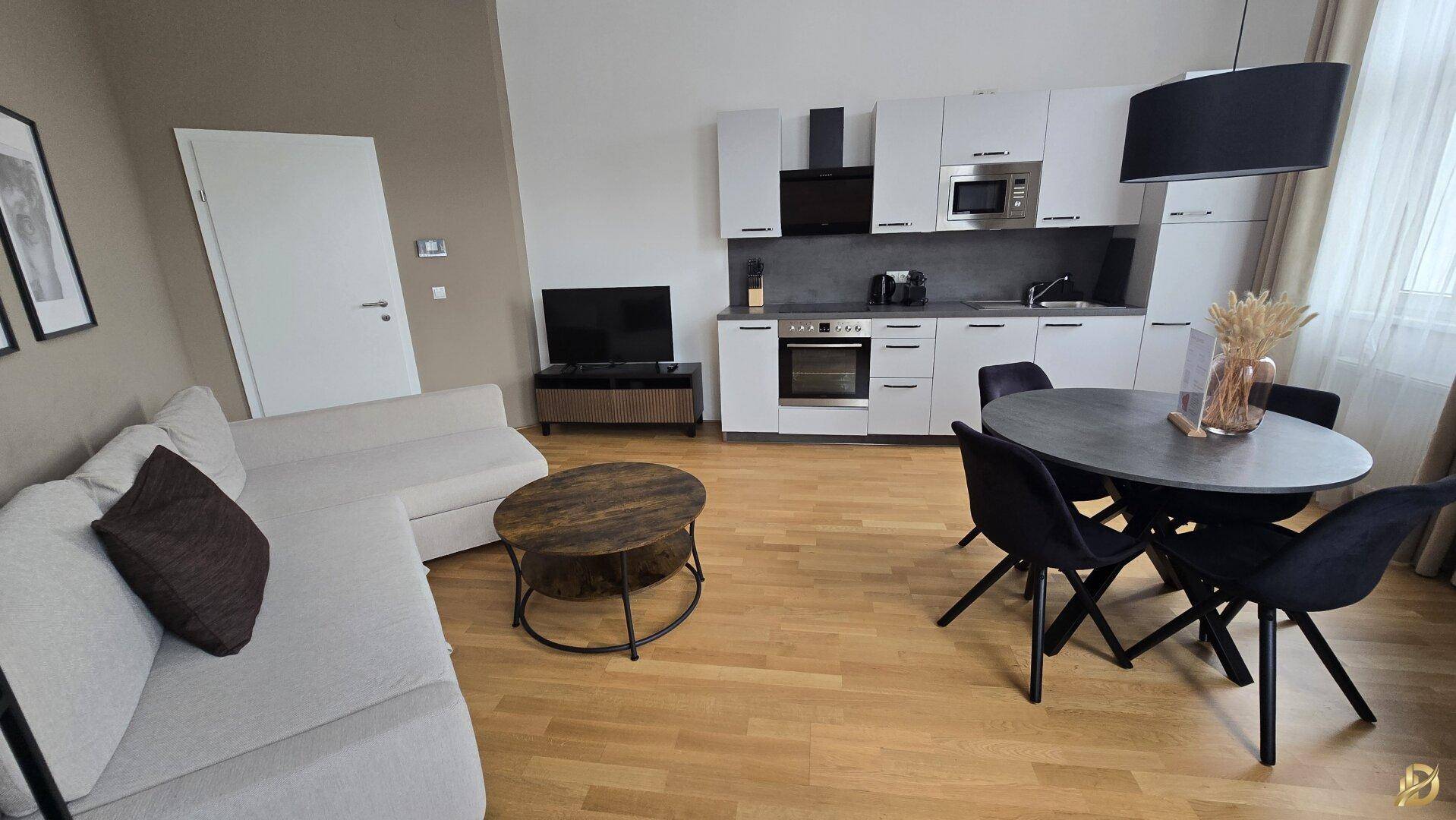 Gemütlicher Wohnbereich mit hellem Sofa, Couchtisch und integrierter, moderner Küche auf Parkettboden.