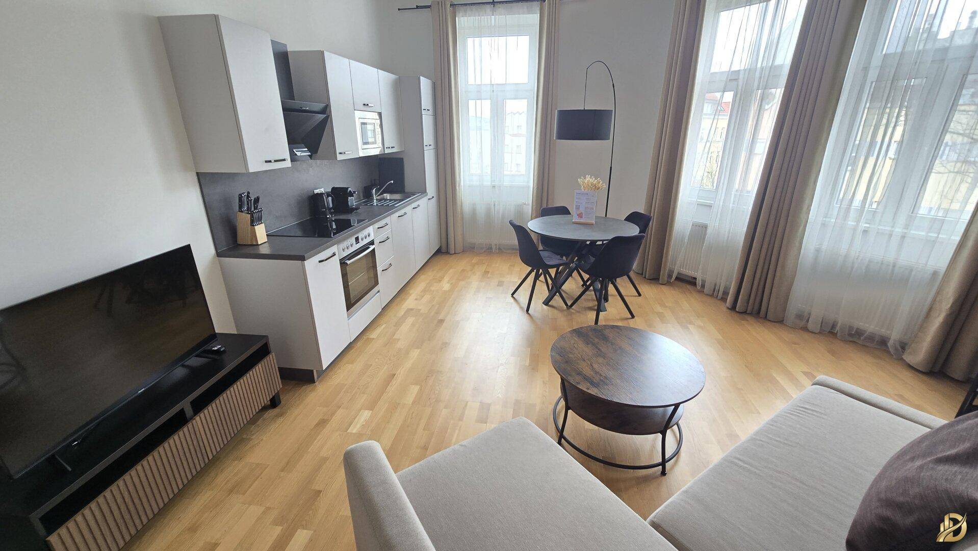 Offener Wohnbereich mit integrierter Küche, Essplatz, Sofa und Fernseher, alles auf hellem Parkettboden.
