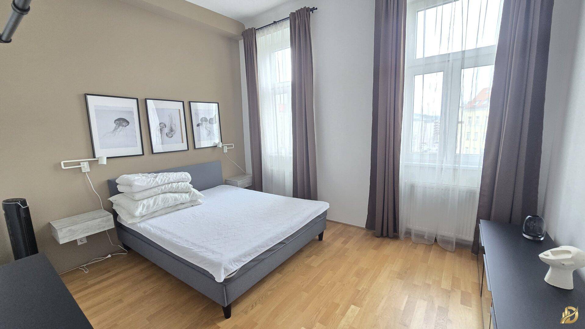 Schlafzimmer mit beigem Bett, Parkettboden und hohen Fenstern, die viel Tageslicht hereinlassen.