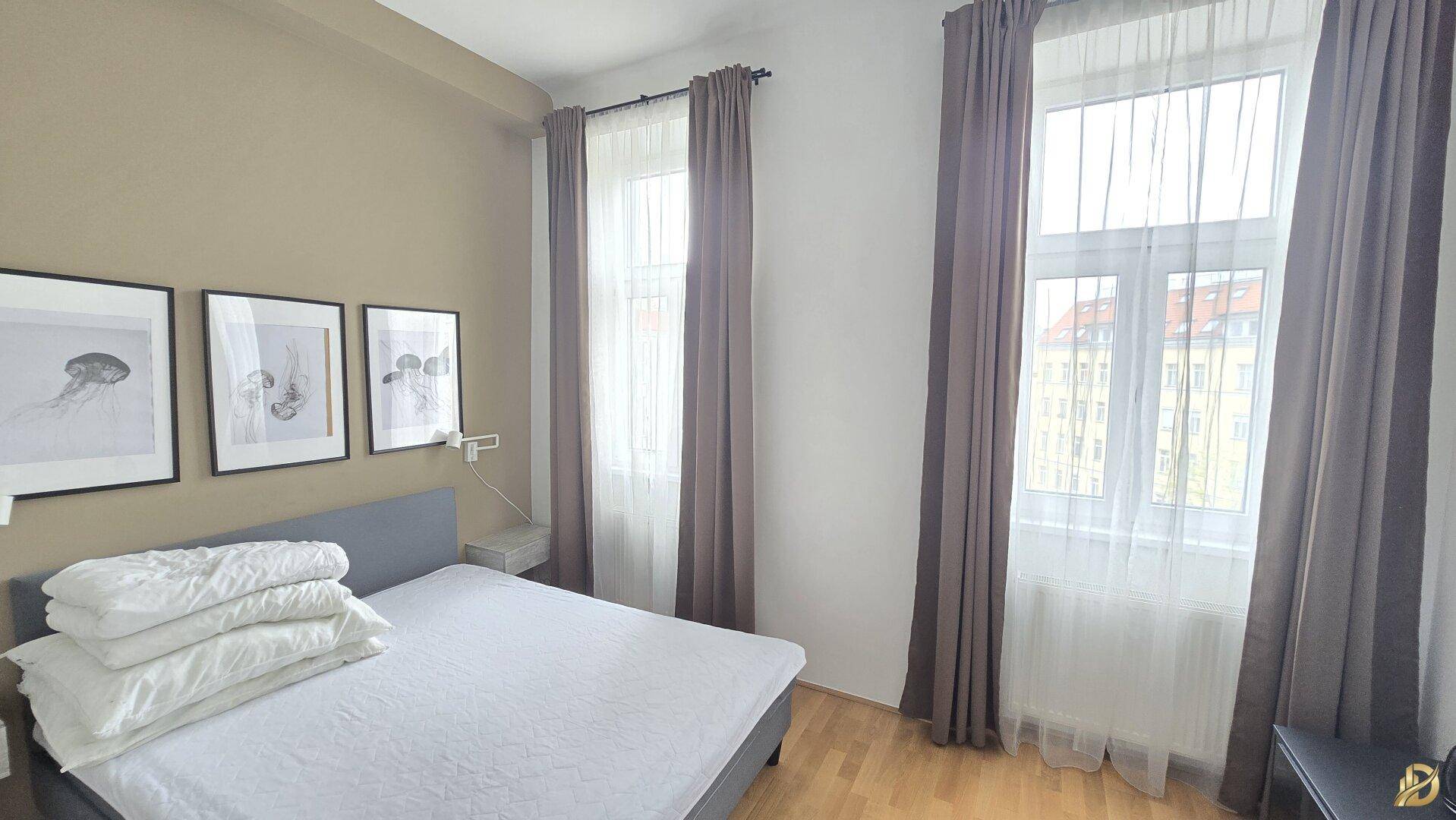 Helles Schlafzimmer mit Doppelbett, Parkettboden und hohen Fenstern mit Vorhängen.