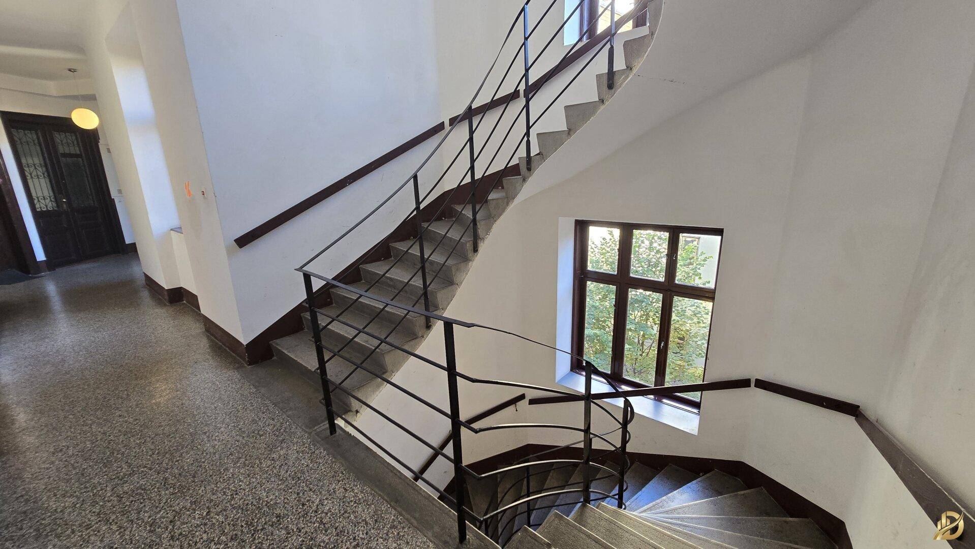 Historisches Treppenhaus mit geschwungenem Geländer und großen Fenstern, die den Bereich erhellen.