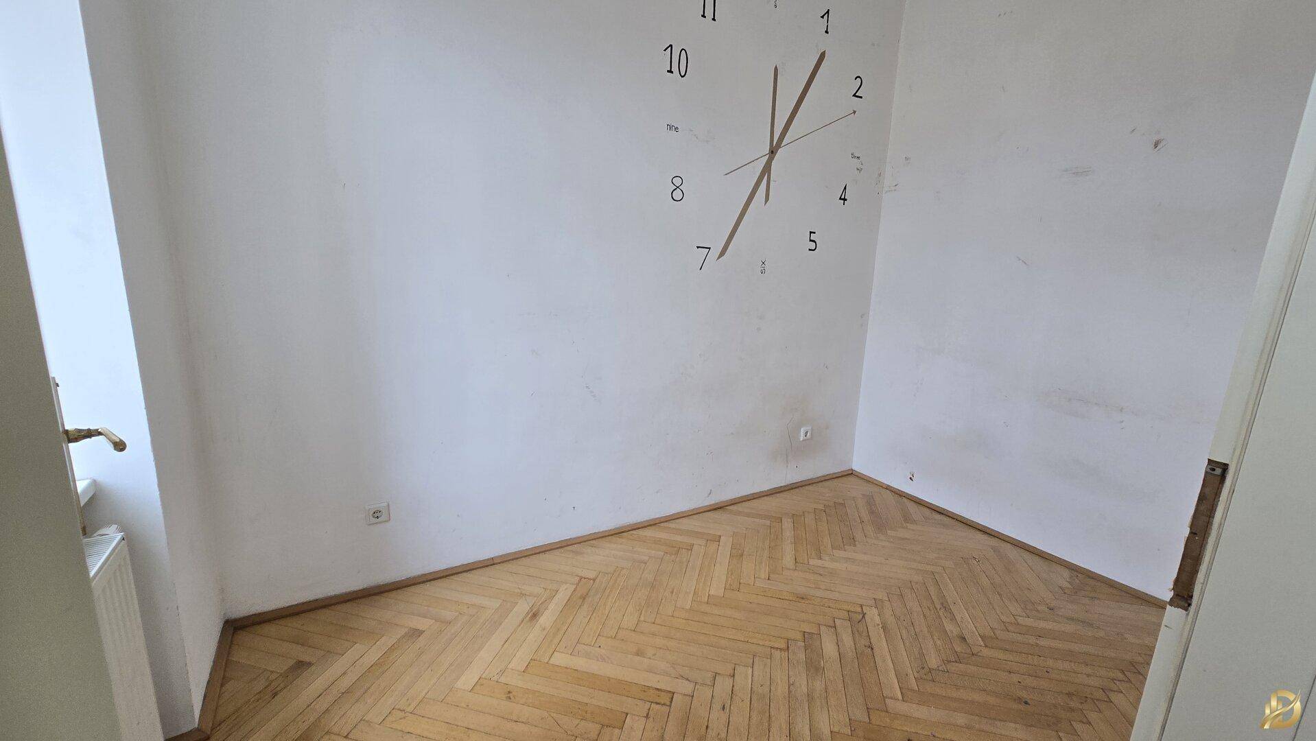 Helles Zimmer mit Fischgrätparkett und einer dekorativen Wanduhr, ideal als Schlaf- oder Arbeitszimmer.