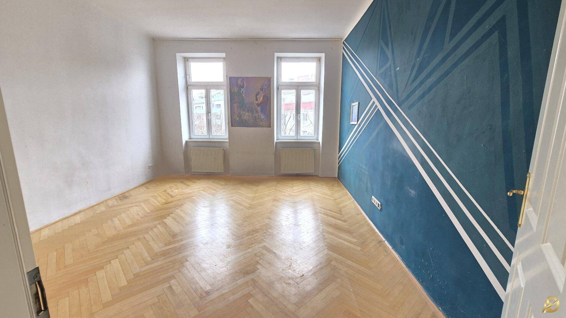 Geräumiges Zimmer mit hellem Fischgrätparkett und einer auffälligen blauen Akzentwand.