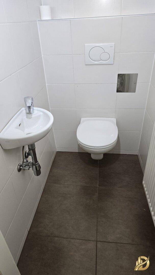 Separates WC mit wandhängender Toilette, kleinem Waschbecken und Fliesenboden.