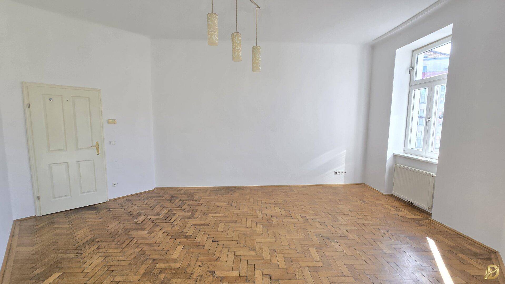 Leeres Zimmer mit Fischgrätparkett, einer Tür und einem Fenster, das viel Licht spendet.