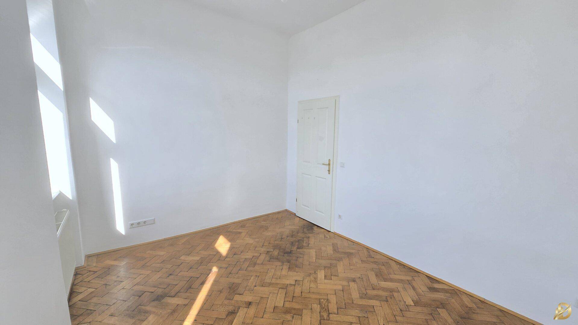 Sonniger Raum mit Fischgrätparkett und einer Tür, die den Zugang zum Zimmer bietet.