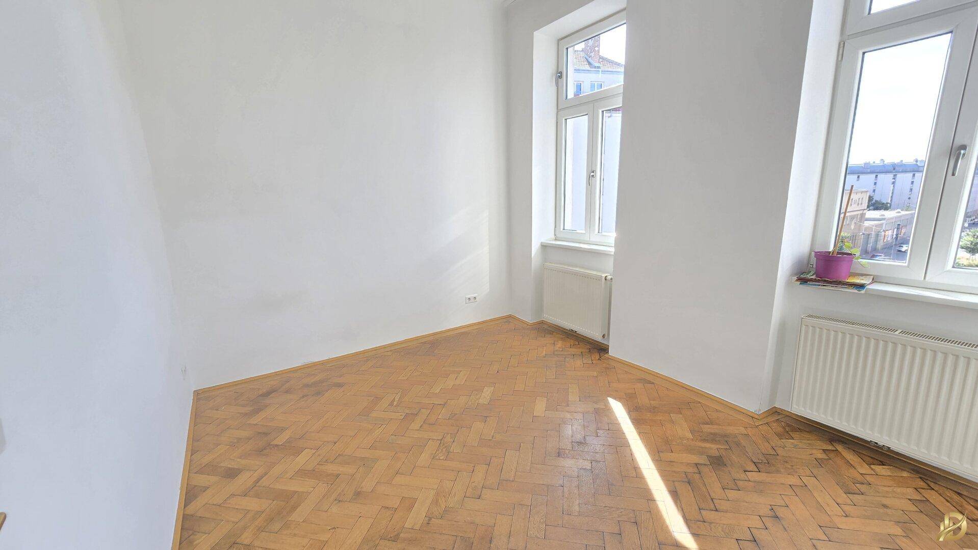 Helles Zimmer mit Parkettboden und zwei Fenstern, die einen sonnigen Ausblick ermöglichen.