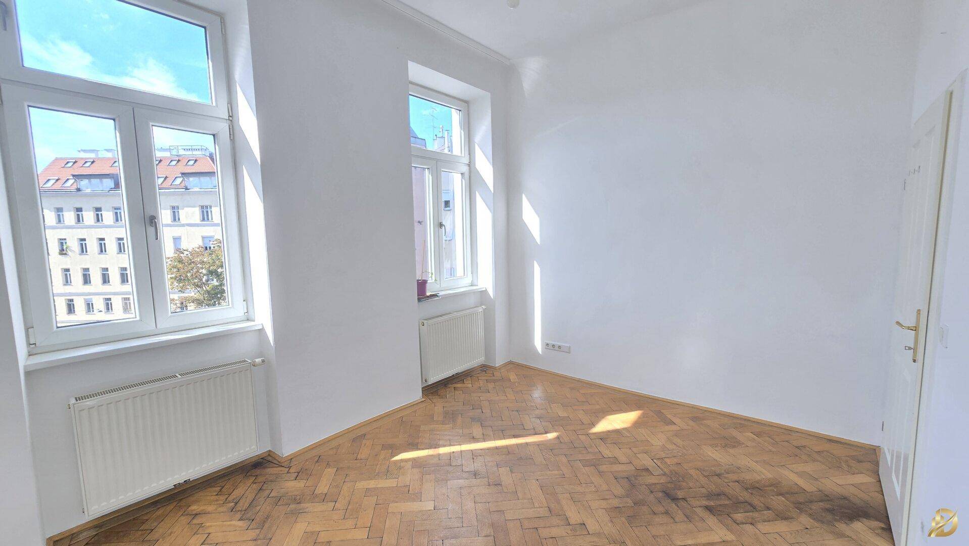 Lichtdurchflutetes Zimmer mit Parkettboden und zwei Fenstern, die einen Blick nach draußen bieten.