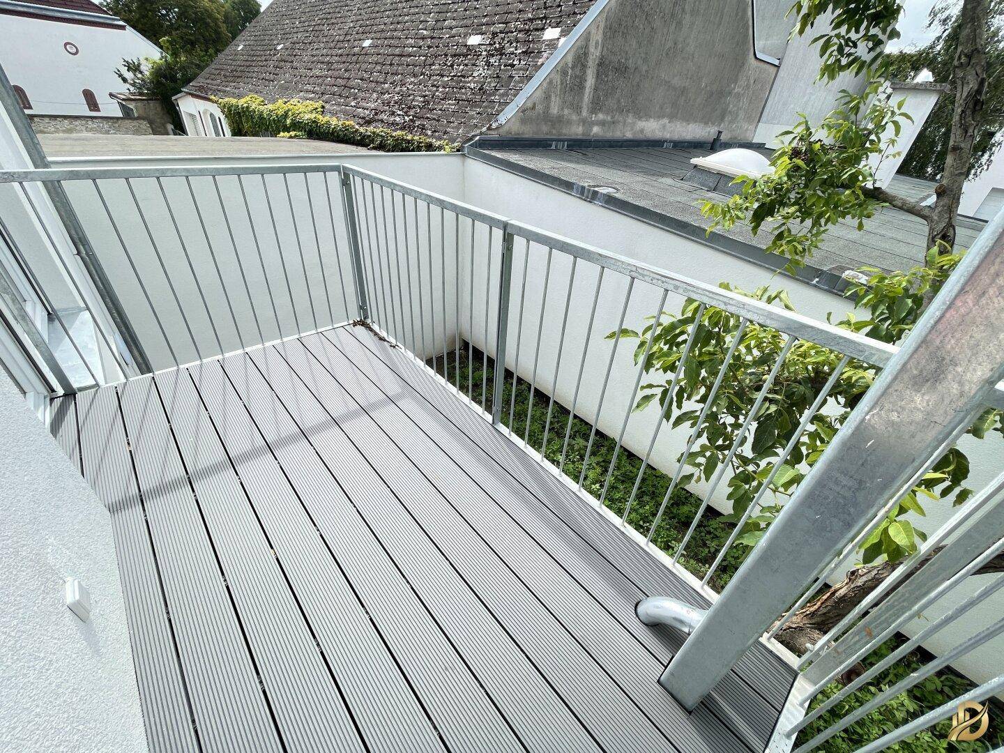 Balkon mit grauem Dielenboden und Metallgeländer, umgeben von etwas Grün.
