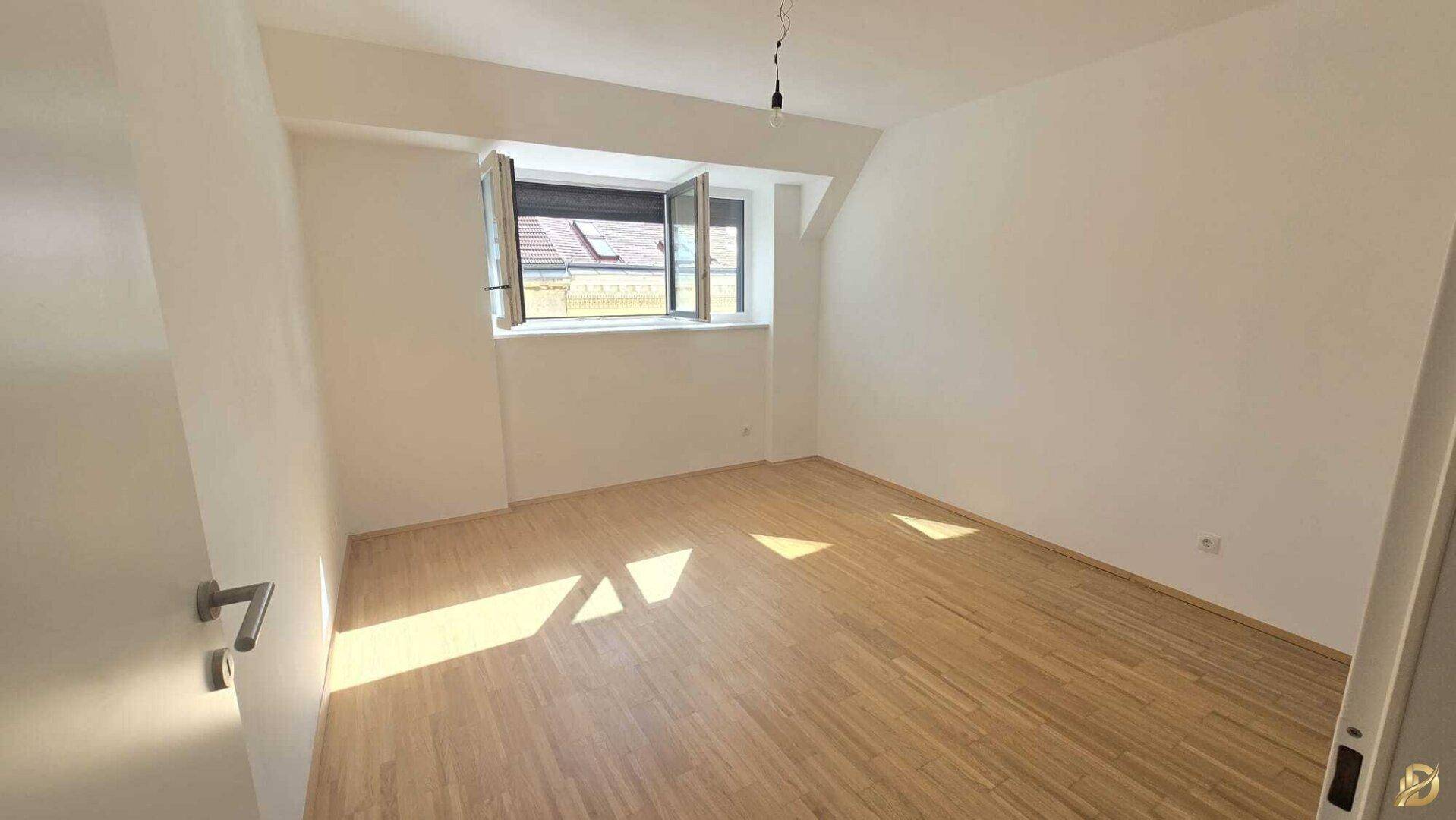 Helles Zimmer mit Holzboden und Fenster, das viel Tageslicht hereinlässt und eine angenehme Atmosphäre schafft.