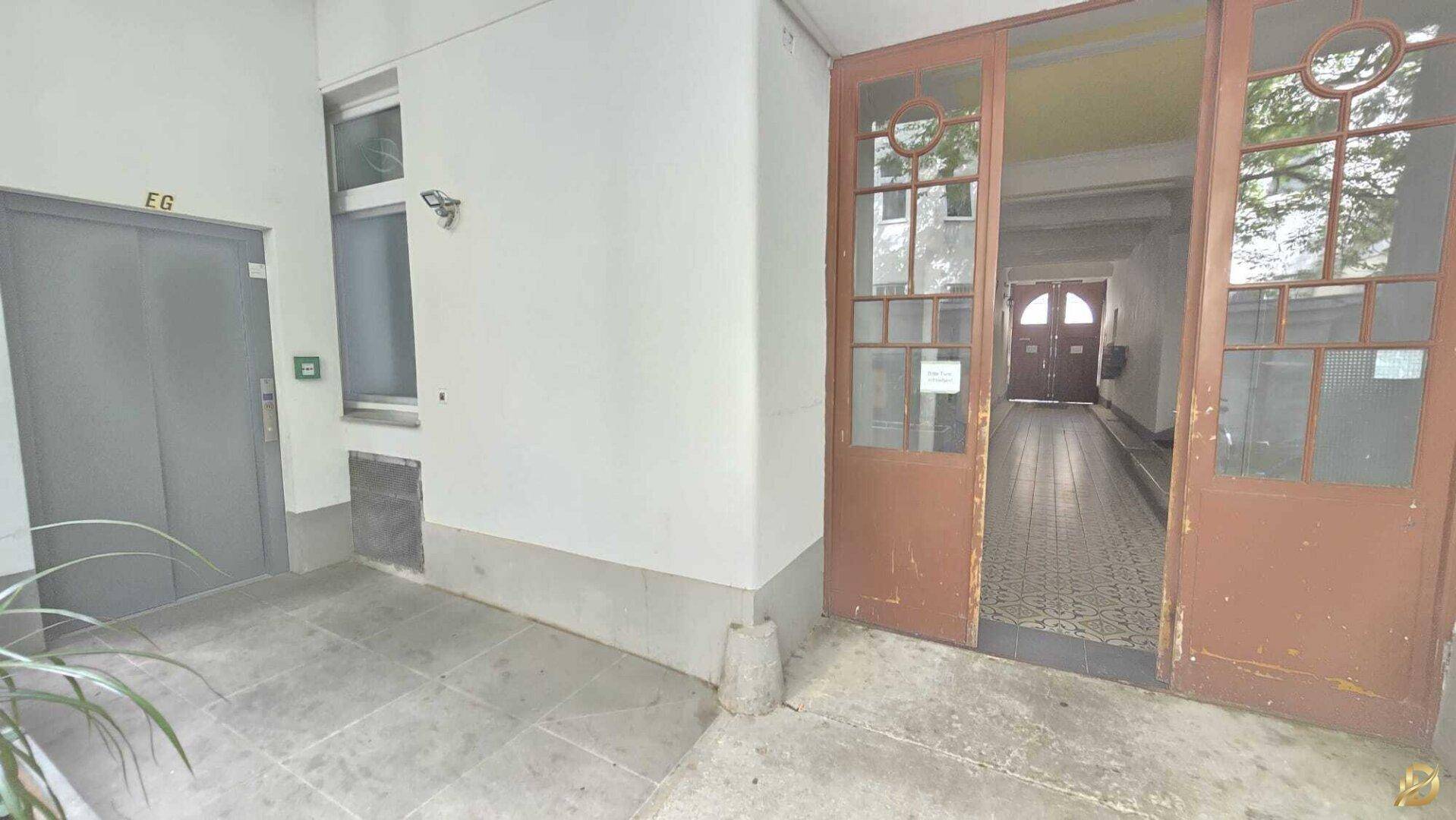 Eingangsbereich eines Gebäudes mit Aufzug und dekorativer Glastür, die zum Flur führt.