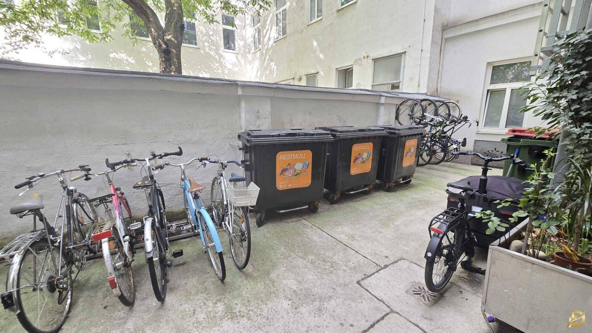 Außenbereich mit Fahrradständern, Mülltonnen und Bäumen im Hintergrund, praktisch und funktional gestaltet.