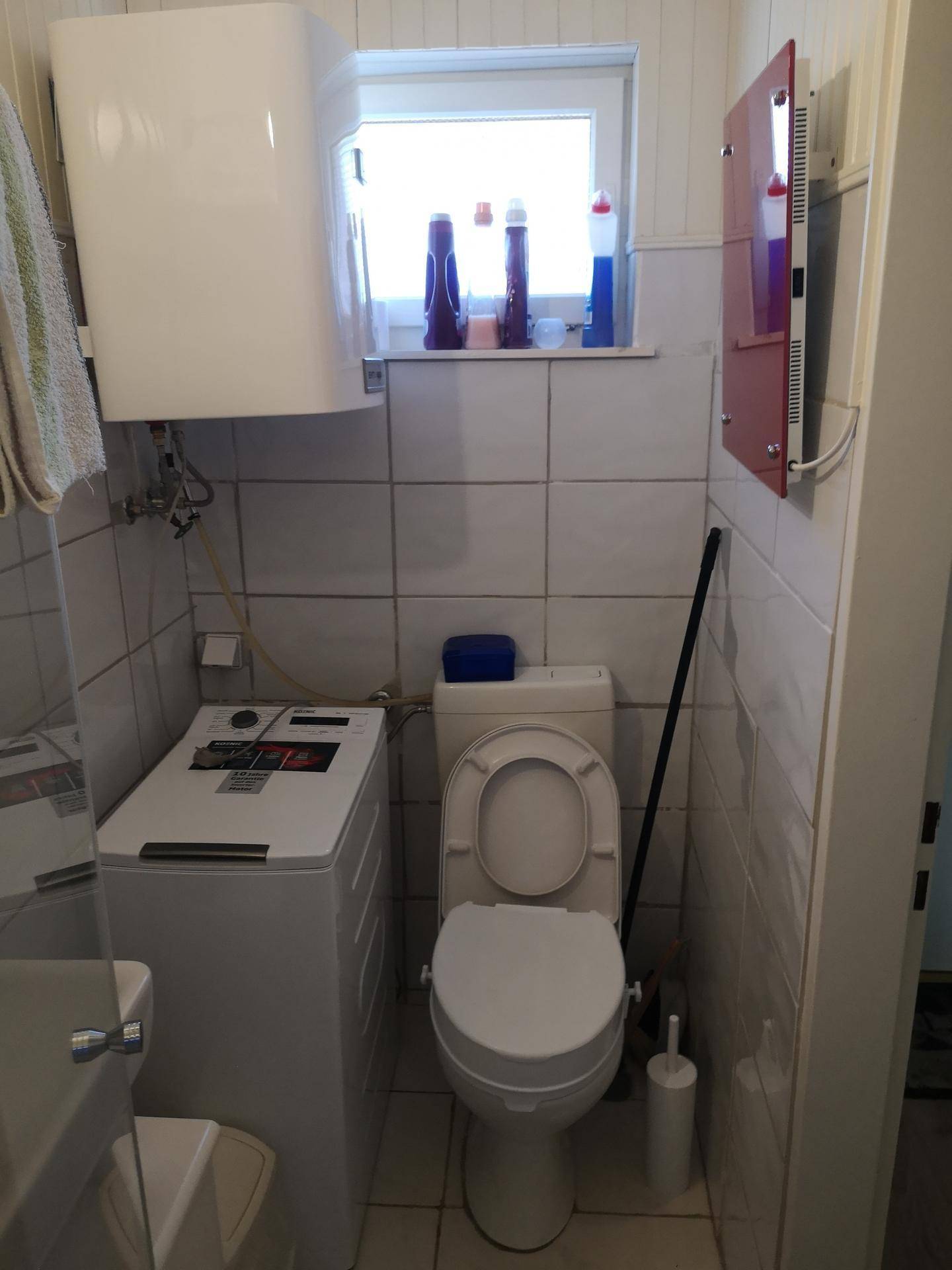 Kleines WC mit Toilette, Waschmaschine und Fenster, komplett gefliest für einfache Pflege.
