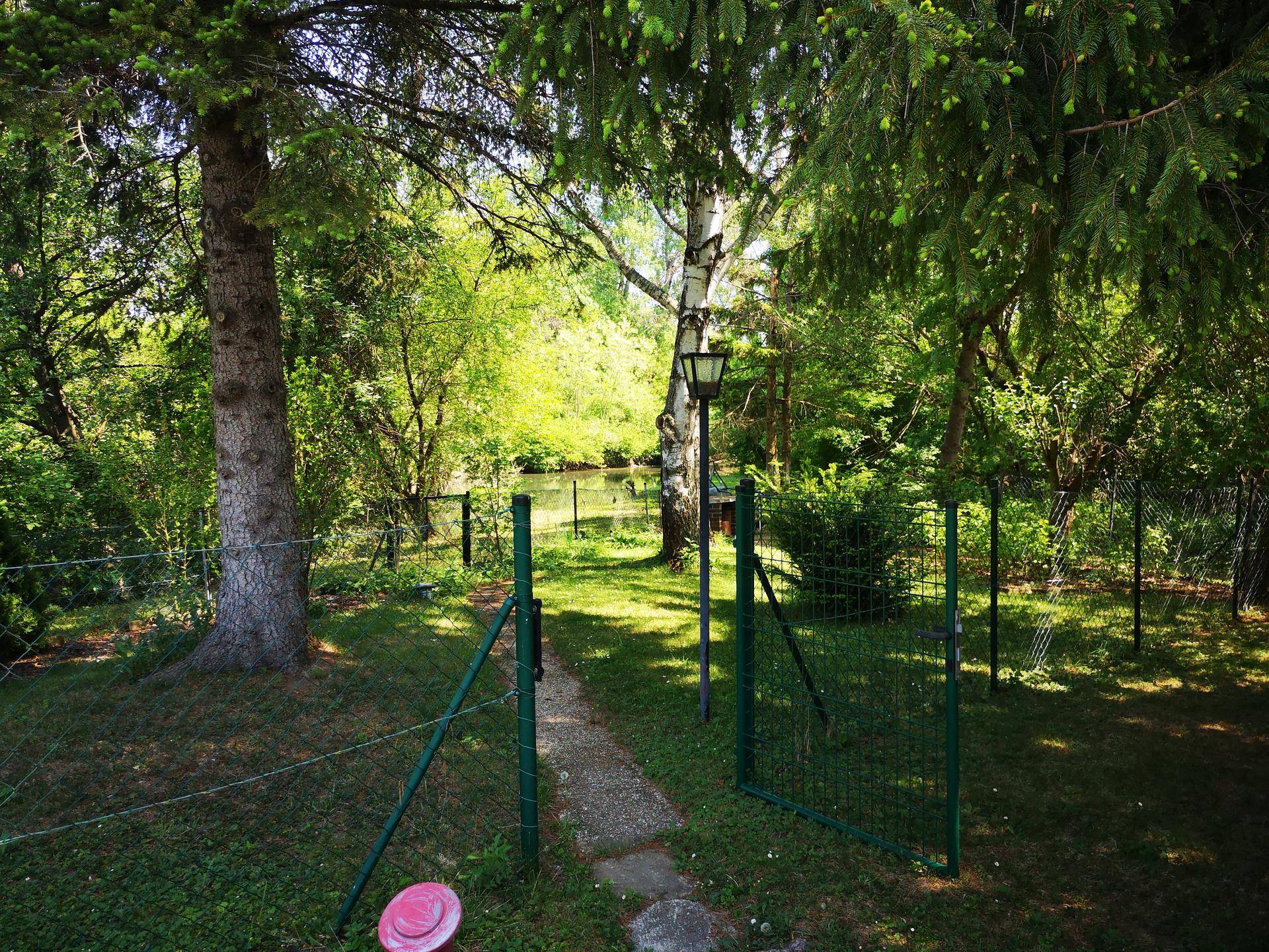 Grüner Garten mit Zaun und Tor, einem Pfad und Blick auf ein Gewässer durch die Bäume.