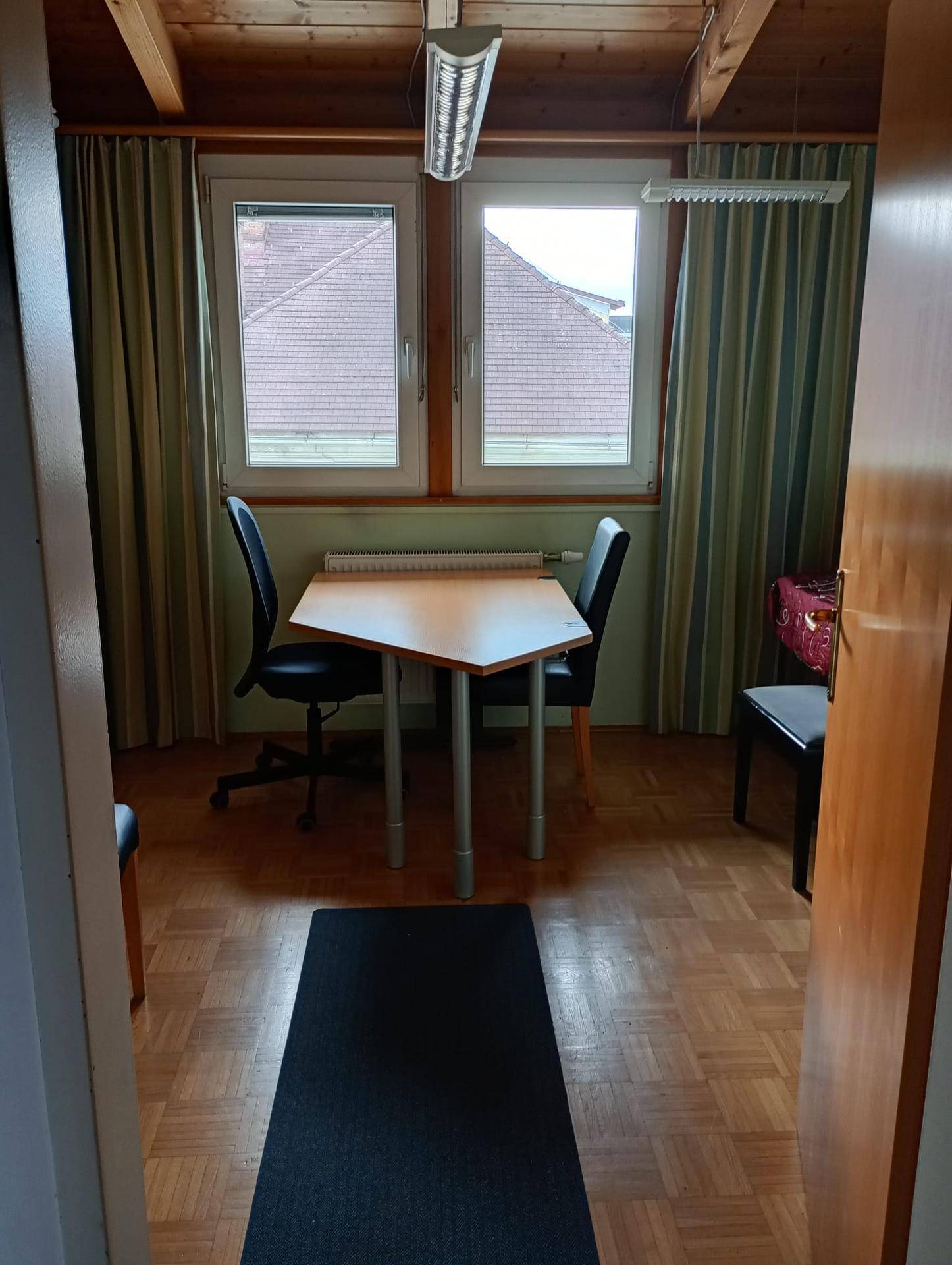 Kleiner Arbeitsbereich mit Schreibtisch, Bürostühlen und Blick auf Dächer durch die Fenster.