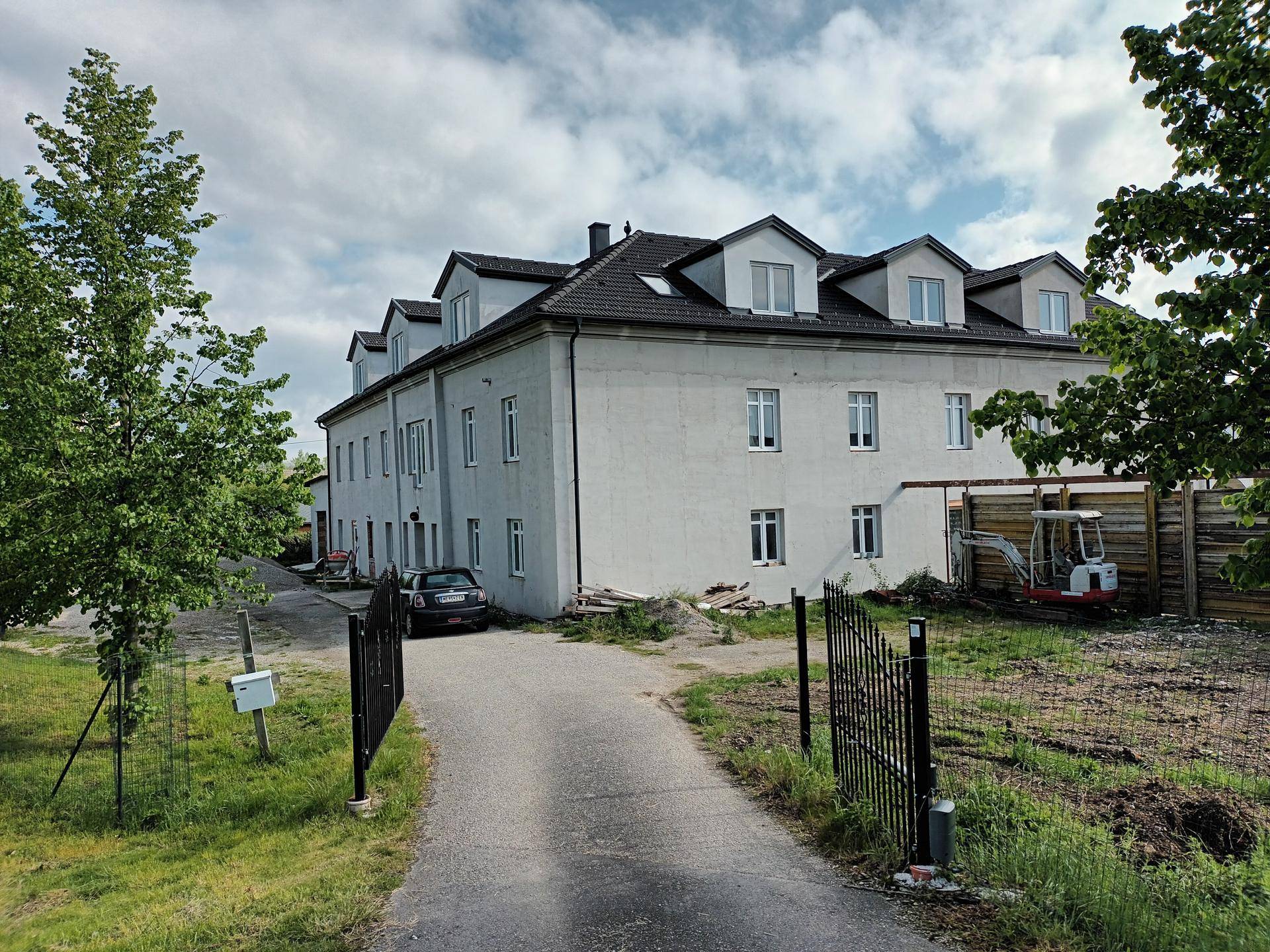 Hauptgebäude mit Zufahrt, offenem Tor und Baumaschinen im Hintergrund, umgeben von Grün.