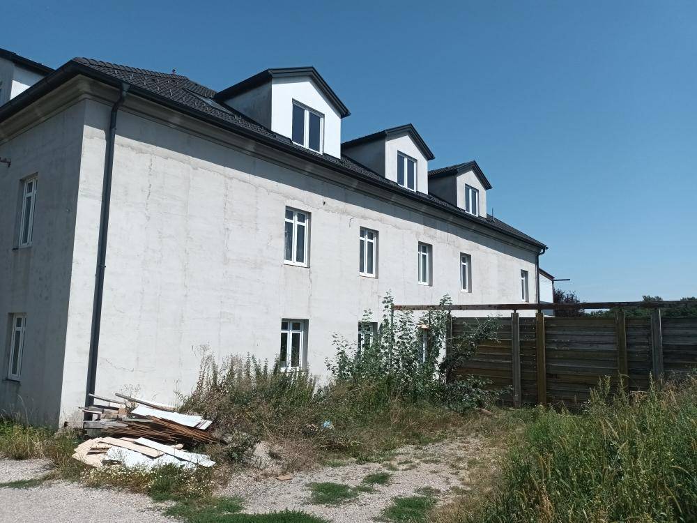 Seitenansicht des Gebäudes mit Zaun und Baumaterialien im Vordergrund, unter klarem Himmel.