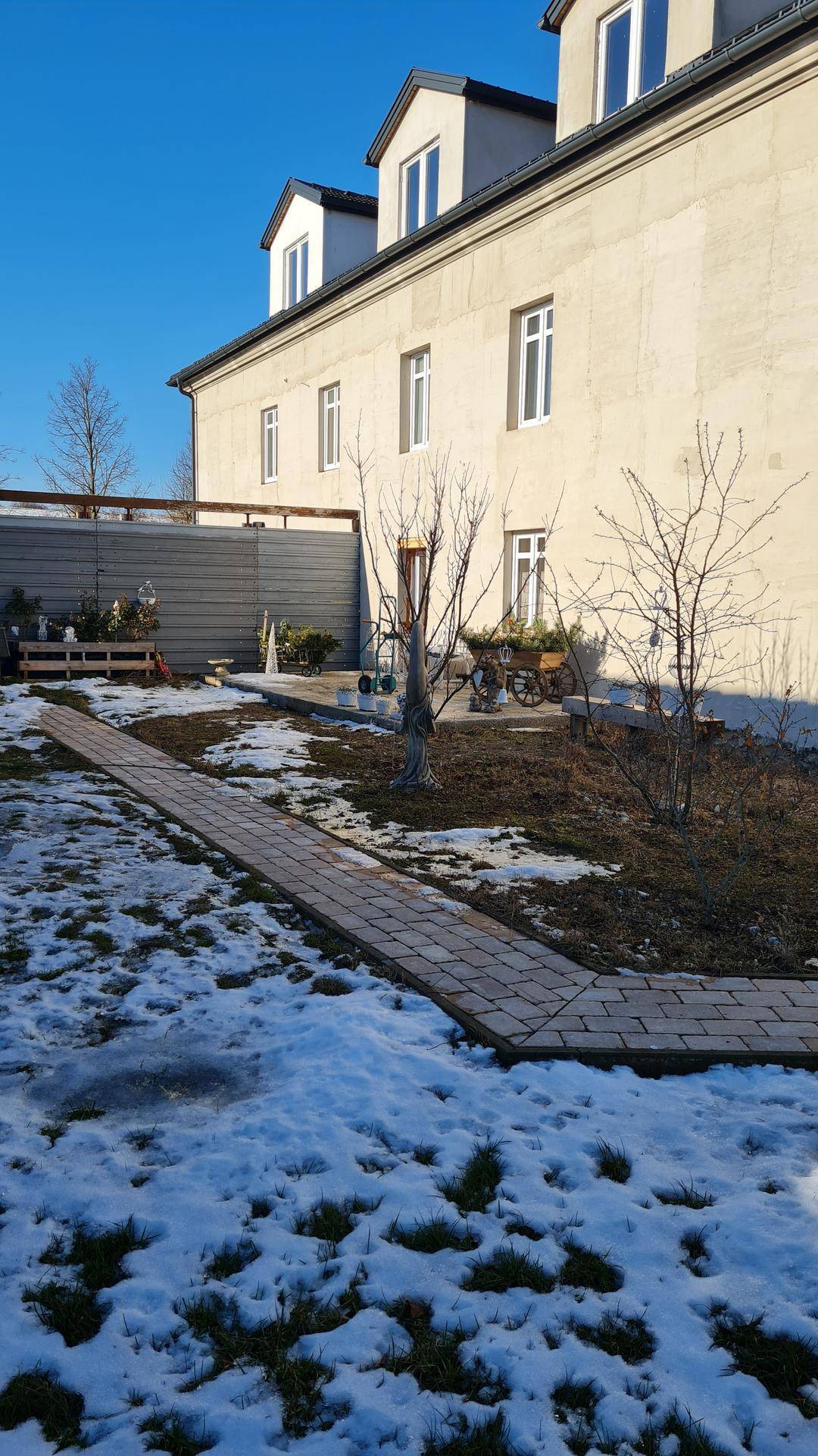 Winterlicher Garten mit verschneitem Rasen, gepflastertem Weg und der Gebäudefassade im Sonnenlicht.