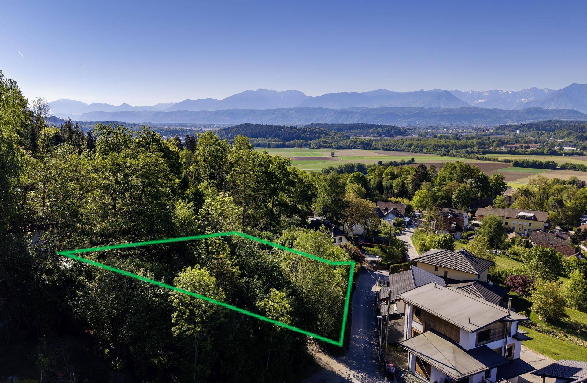 Luftaufnahme eines bewaldeten Grundstücks, umrandet von einer grünen Linie, mit Häusern und Bergen im Hintergrund.