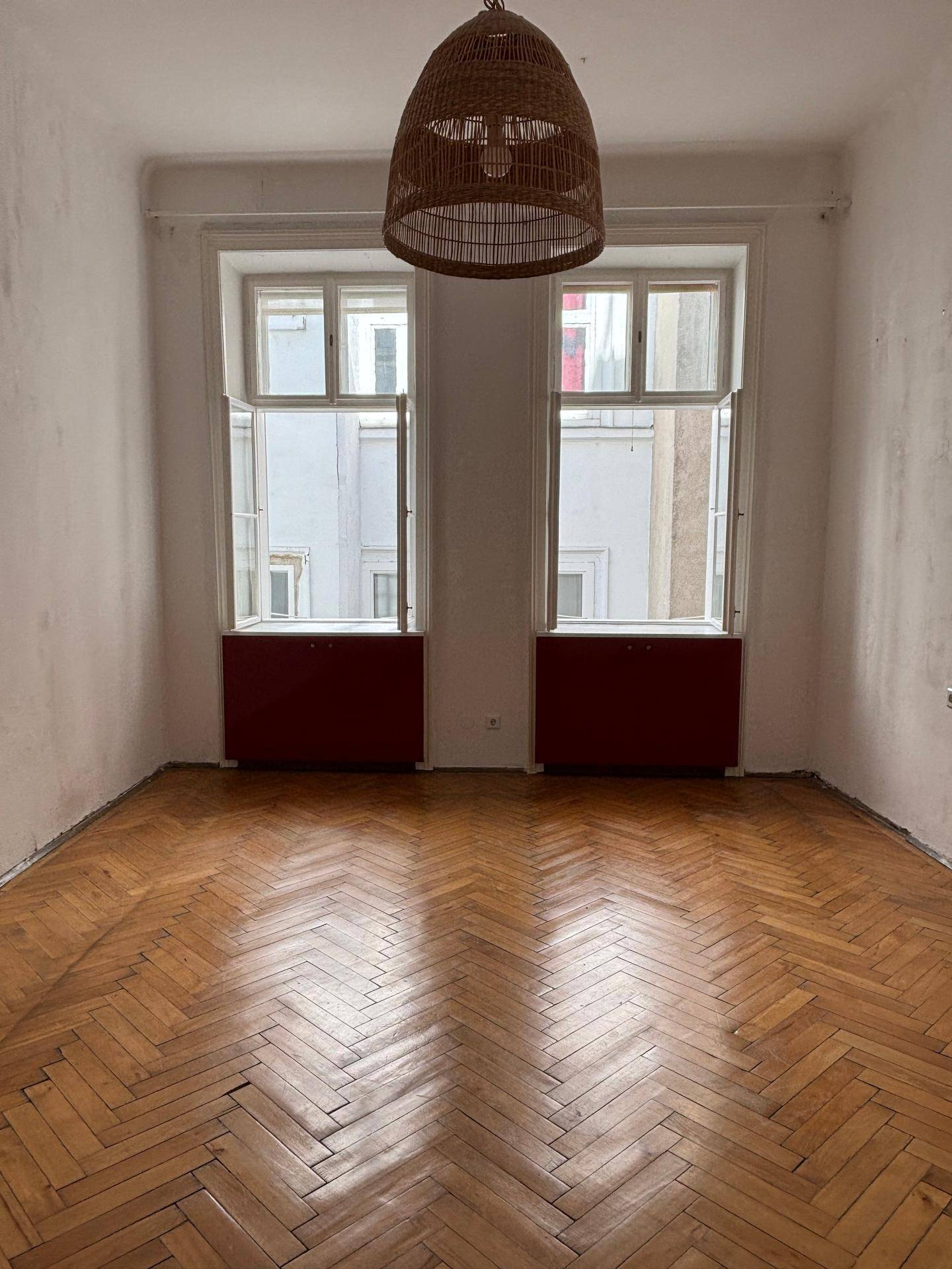 Leeres Zimmer mit Fischgrätparkett, hohen Fenstern und roter Heizungsverkleidung.
