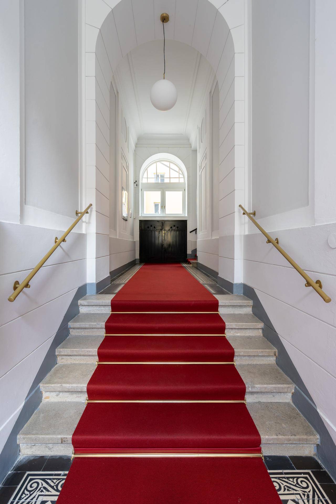 Elegantes Treppenhaus mit rotem Teppich, steinernen Stufen und gewölbter Decke, führt zu einem großen Fenster.