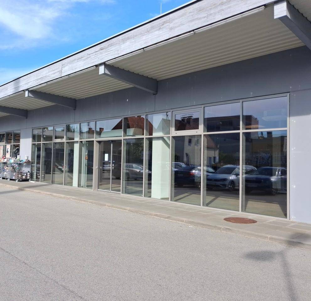 Moderne Geschäftsfassade mit großen Glasfenstern und einem Parkplatz davor unter blauem Himmel.