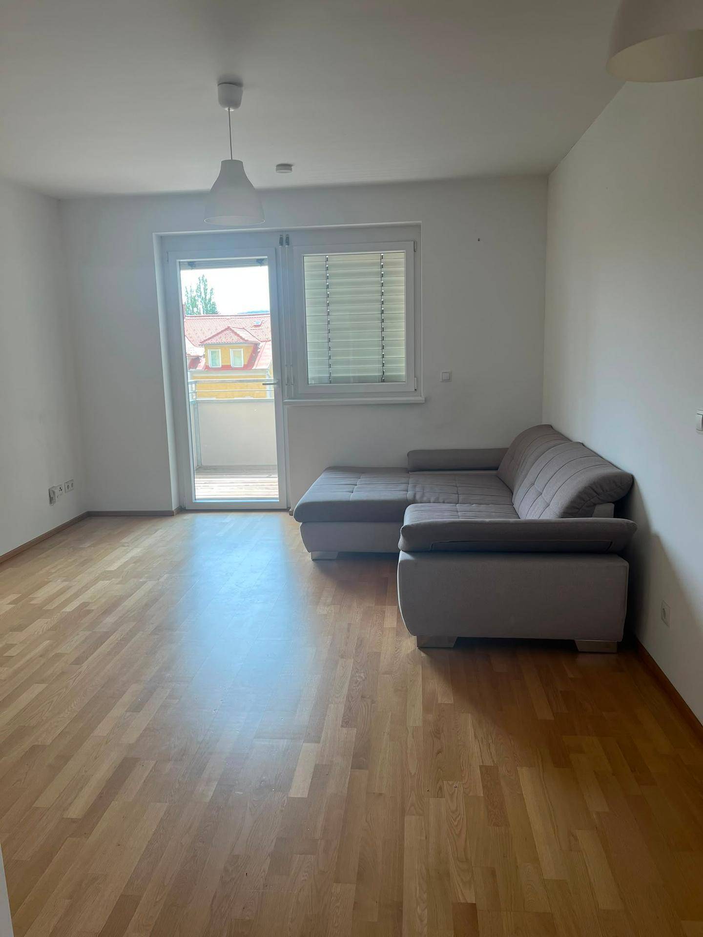 Gemütliches Wohnzimmer mit grauem Ecksofa, Holzboden und direktem Zugang zum Balkon.