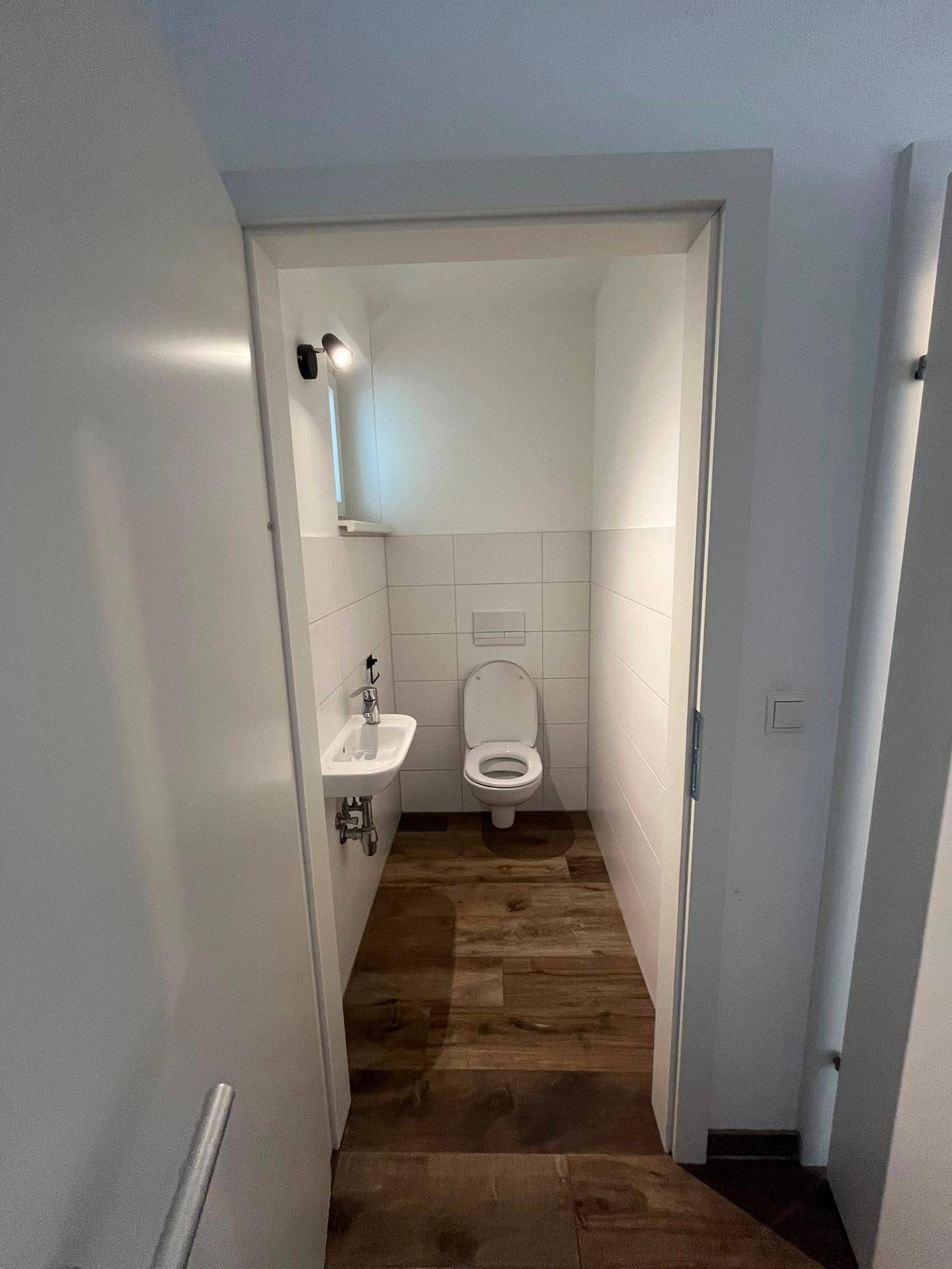Separates Gäste-WC mit wandhängender Toilette, kleinem Waschbecken und Fliesenboden in Holzoptik.