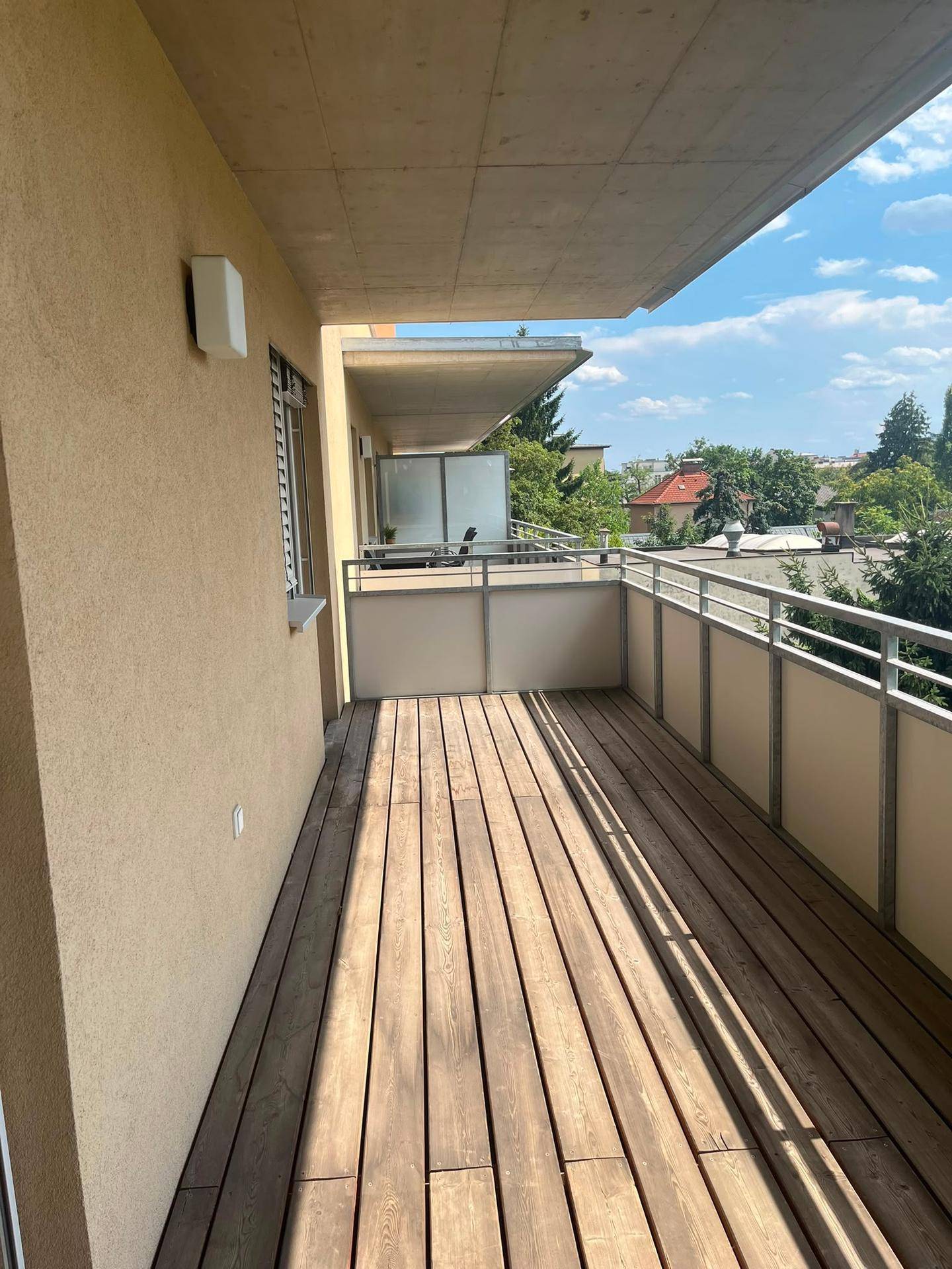 Langer, sonniger Balkon mit Holzdielenboden und Blick ins Grüne sowie auf umliegende Gebäude.