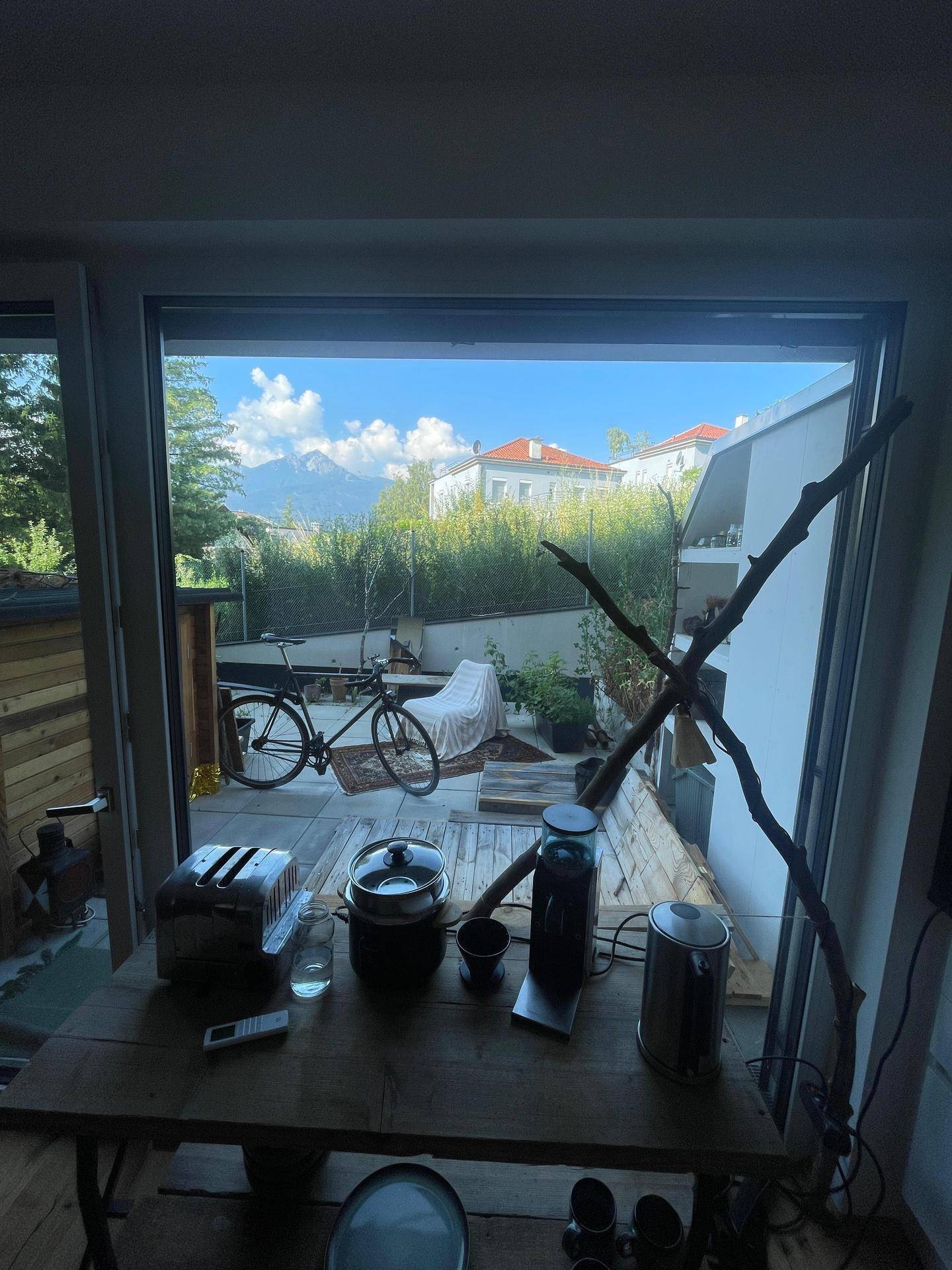 Blick von innen auf die Holzterrasse mit Fahrrad und grüner Umgebung sowie Bergsicht.