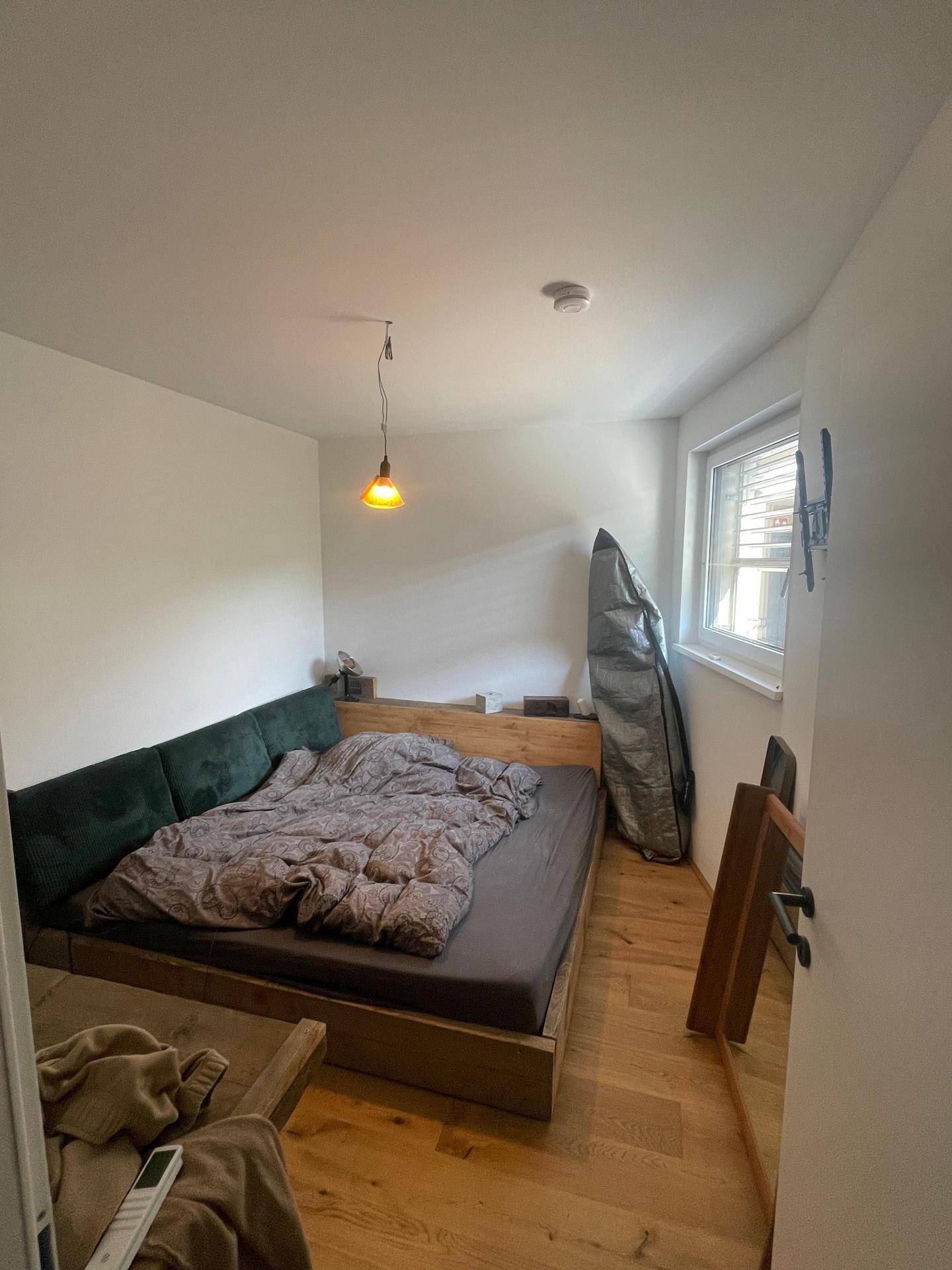 Schlafzimmer mit Holzbettrahmen, dunklem Kopfteil und Fenster für natürliches Licht.