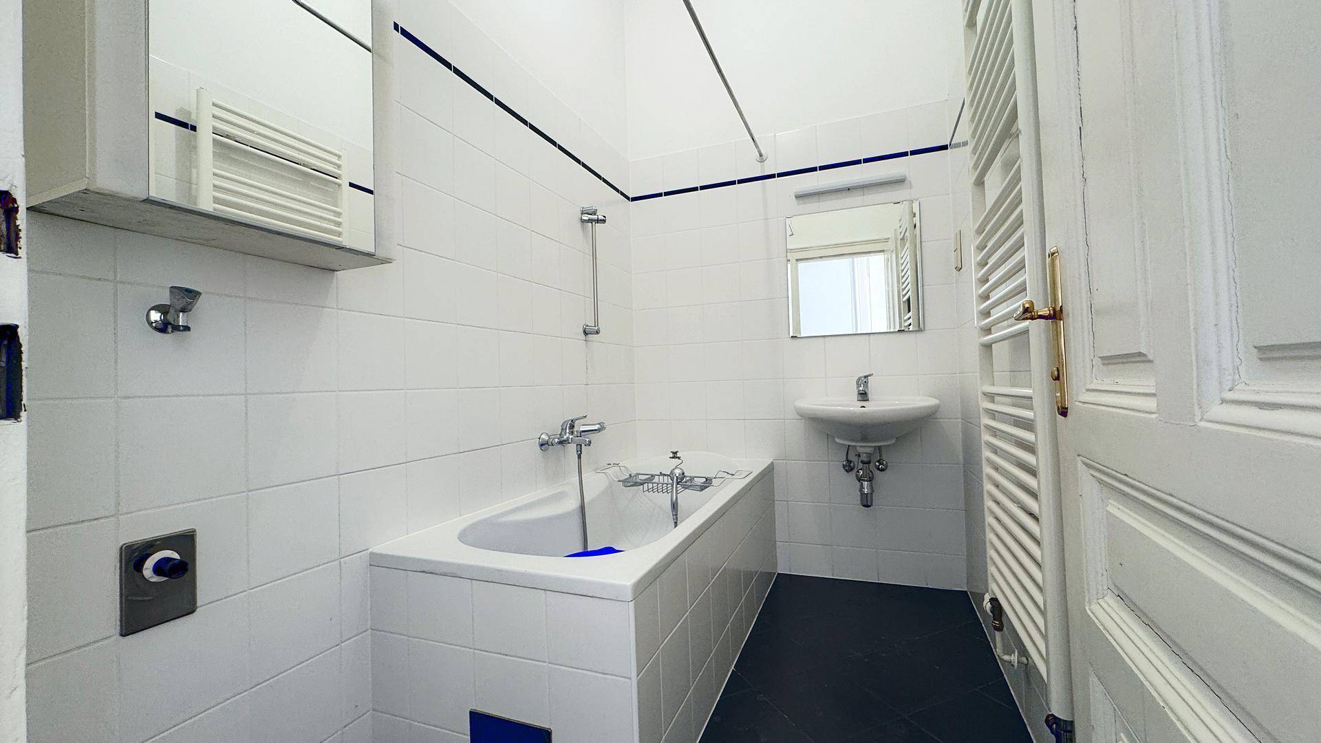 Gefliestes Badezimmer mit Badewanne, Waschbecken und beheiztem Handtuchhalter in weiß-blauer Farbgebung.
