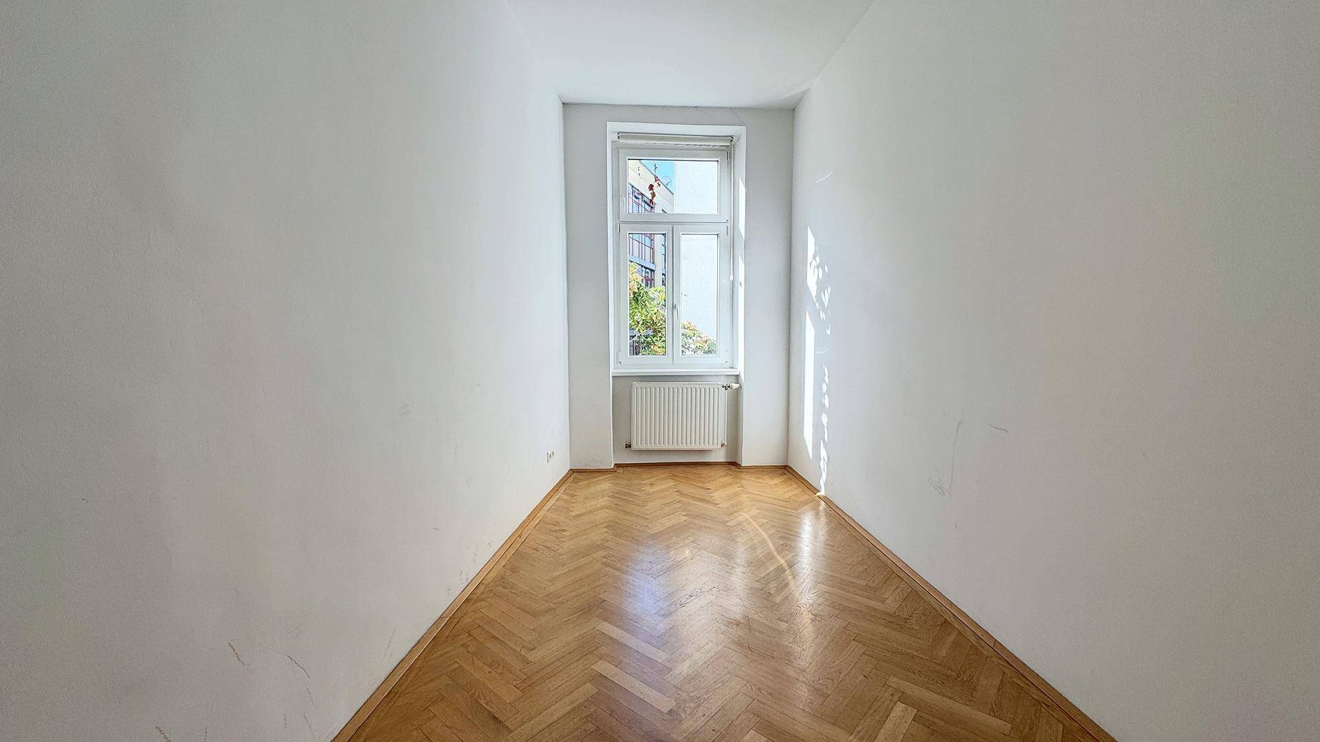 Schmaler, heller Raum mit Fischgrätparkett und einem Fenster am Ende, ideal als Schlaf- oder Arbeitszimmer.