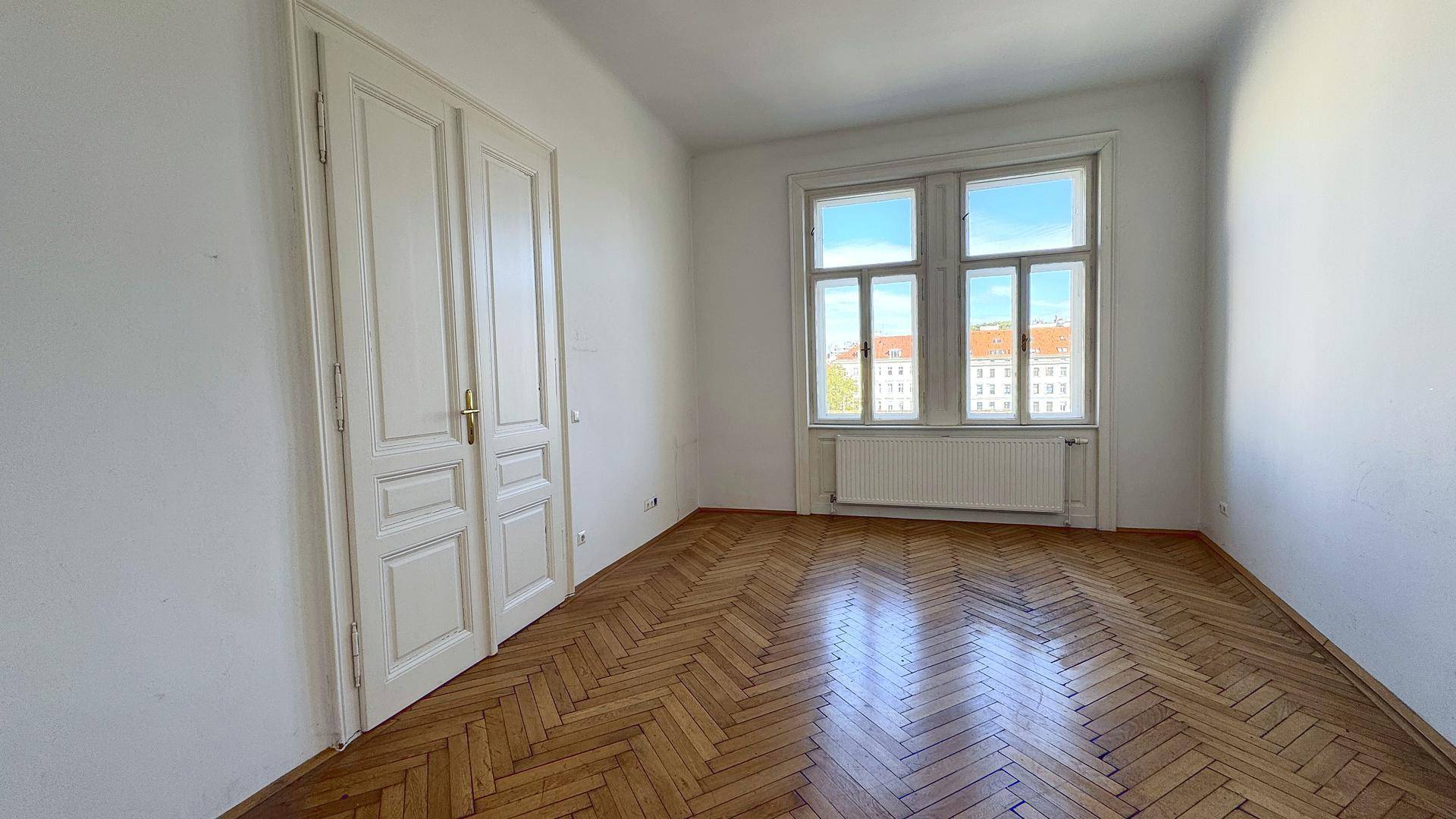 Geräumiges Zimmer mit klassischem Fischgrätparkett und viel Tageslicht durch das große Fenster.