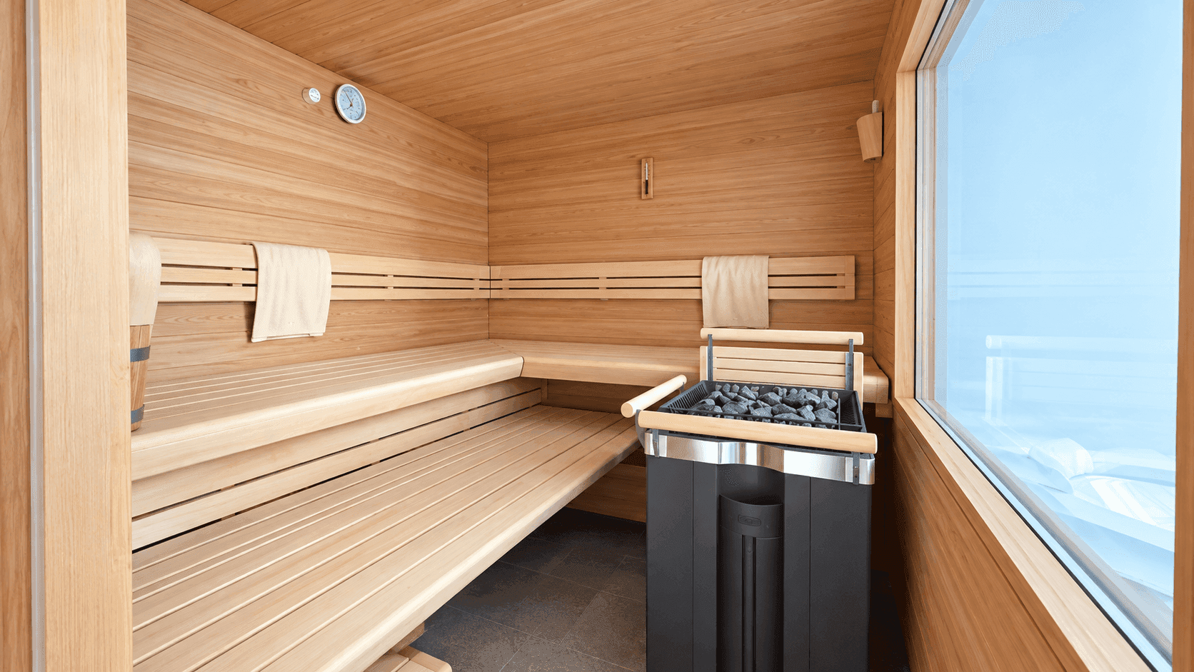Moderne Holzsauna mit breiten Bänken, Saunaofen und großem Fenster für natürlichen Lichteinfall.