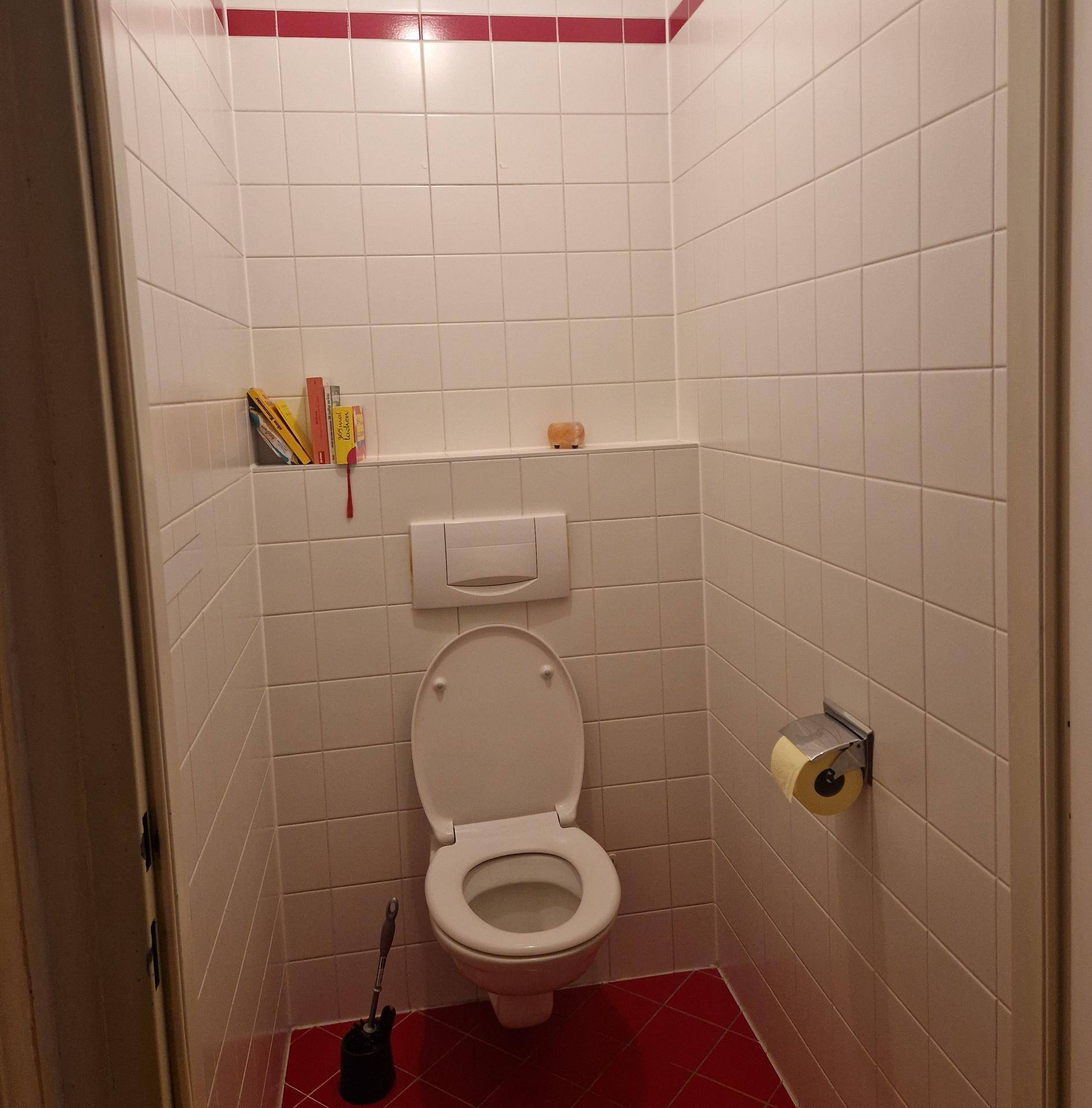 Separates WC mit weißen Wandfliesen, rotem Boden und wandhängender Toilette.