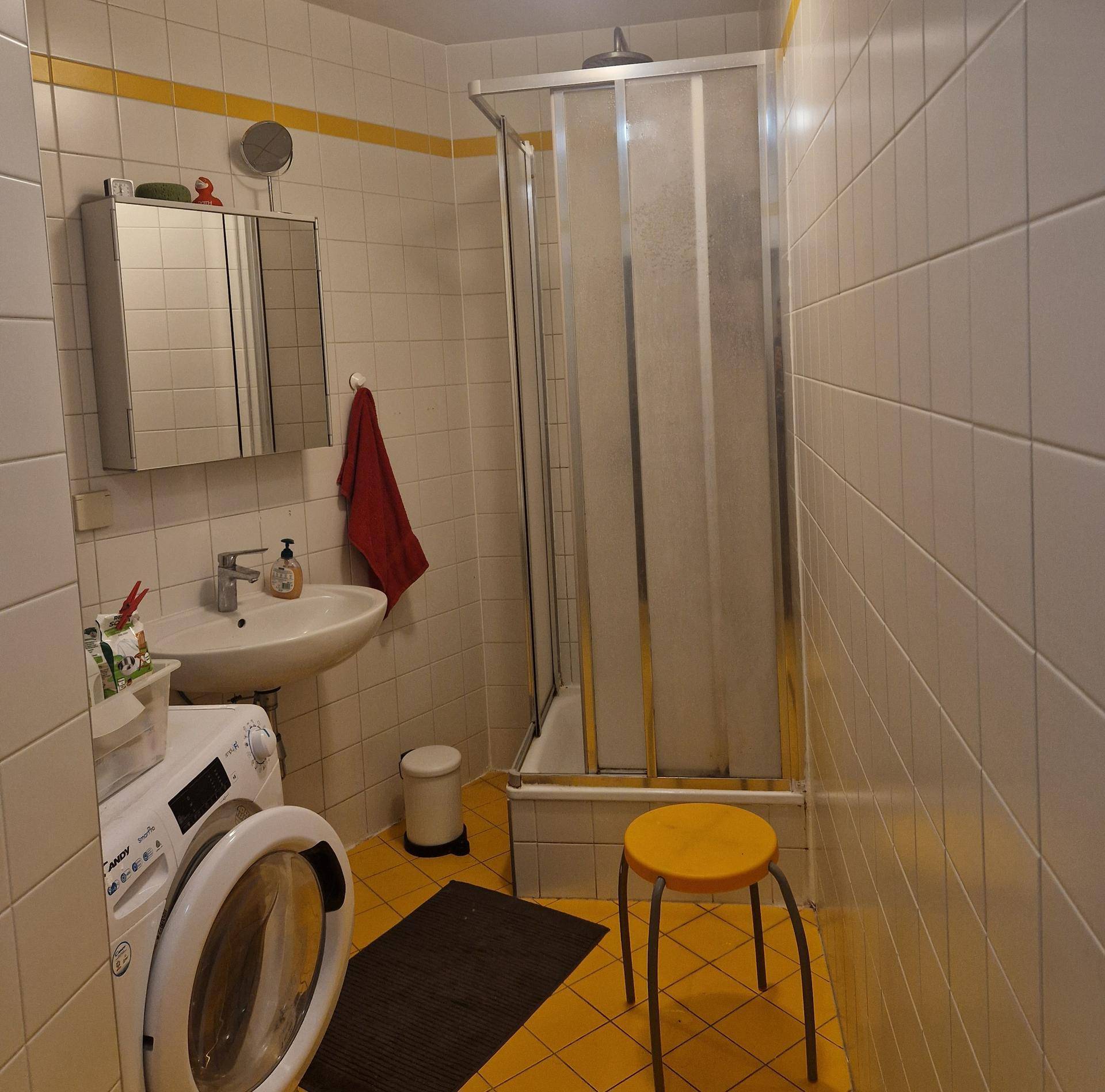 Badezimmer mit weißen Wandfliesen, gelbem Boden, Duschkabine, Waschbecken und Waschmaschine.