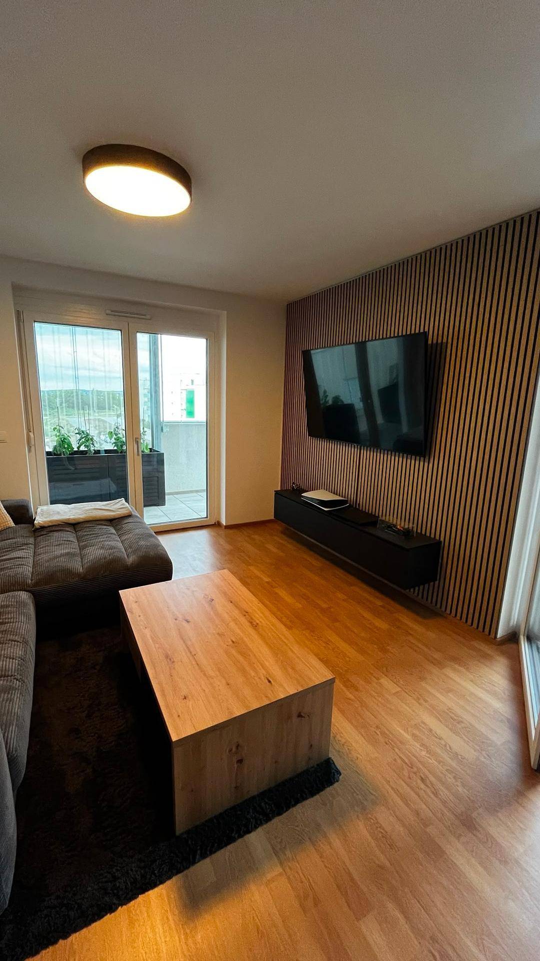 Modernes Wohnzimmer mit TV an einer Lamellenwand, Ecksofa und Balkonzugang, hell und stilvoll.