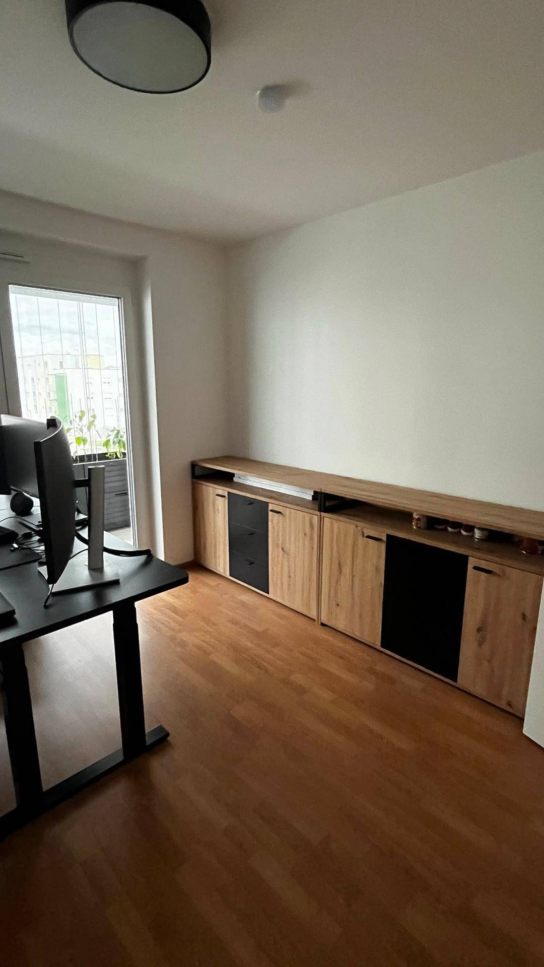 Helles Arbeitszimmer mit Schreibtisch, Monitoren und langen Holzschränken, bietet Balkonzugang.
