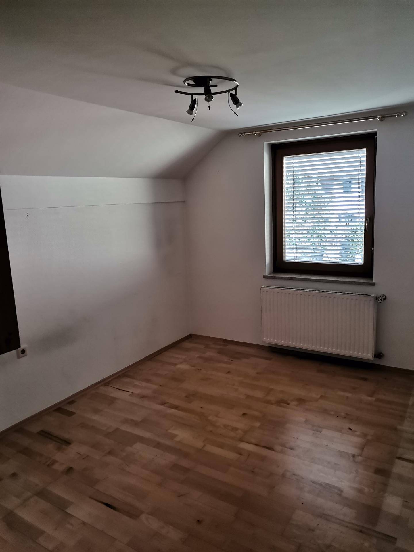 Dachzimmer mit Parkettboden, weiß gestrichenen Wänden und einem Fenster mit Jalousien.