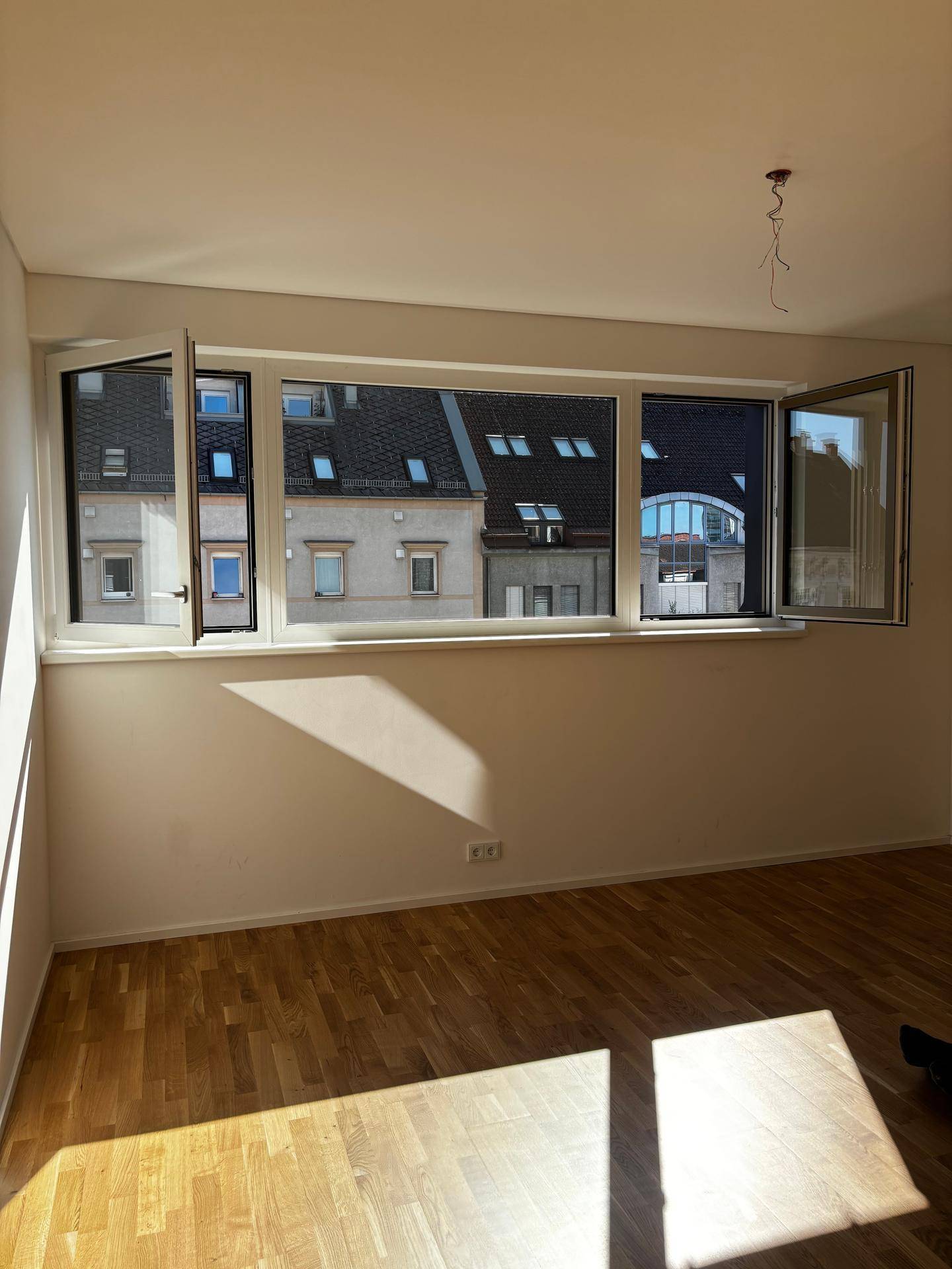 Heller Raum mit Holzboden und großen Fenstern, die einen Blick auf die umliegenden Gebäude bieten.