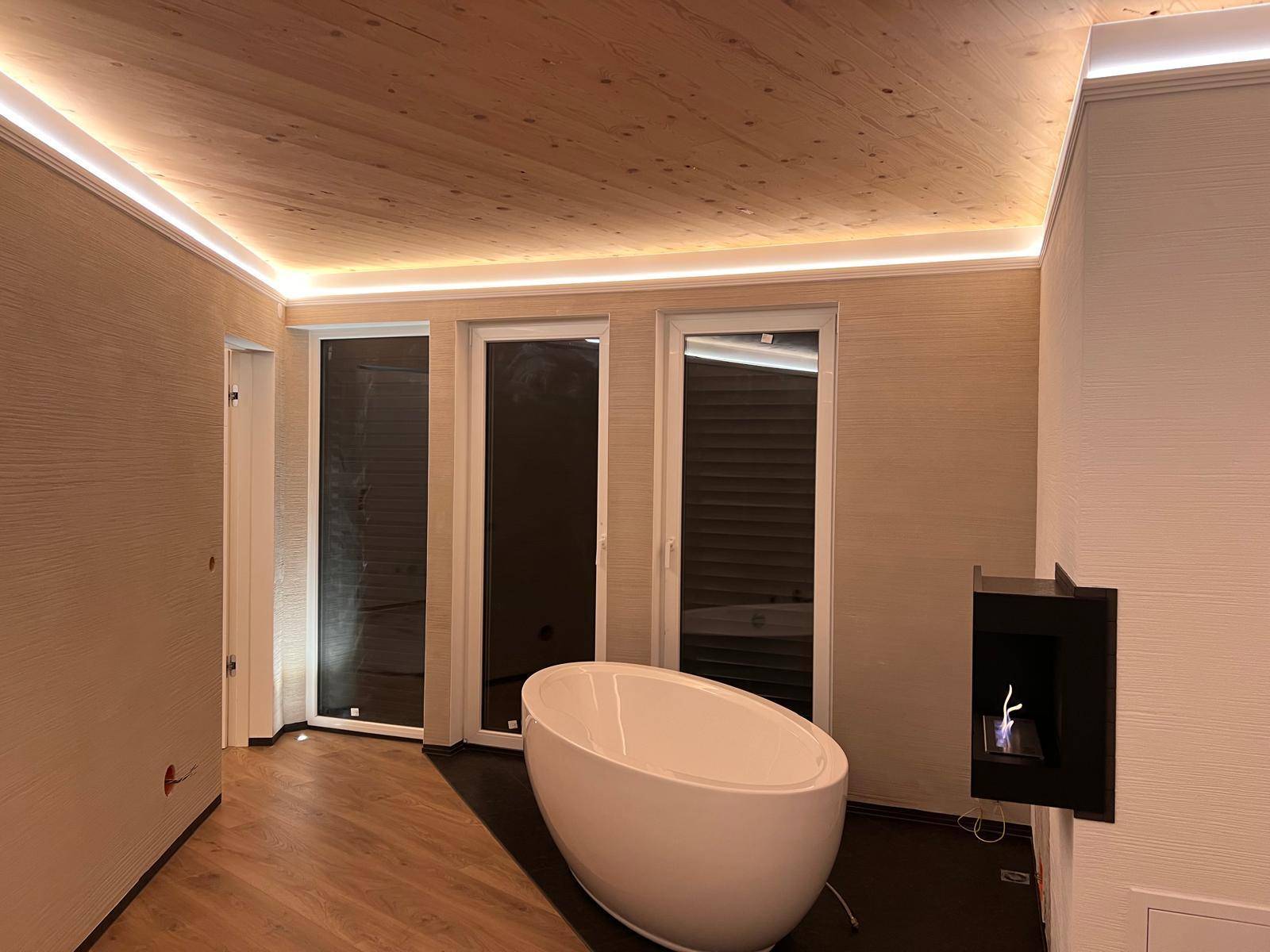 Geräumiger Raum mit Holzboden, freistehender Badewanne und Kamin, umgeben von Fenstern.