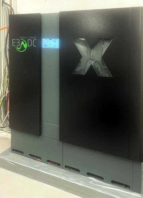 Schwarzes Energie-Speichersystem mit grünem Logo und digitaler Anzeige in einem Technikraum.