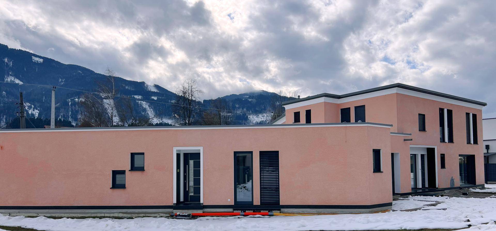 Modernes Haus mit rosafarbener Fassade und dunklen Fenstern in winterlicher Umgebung.