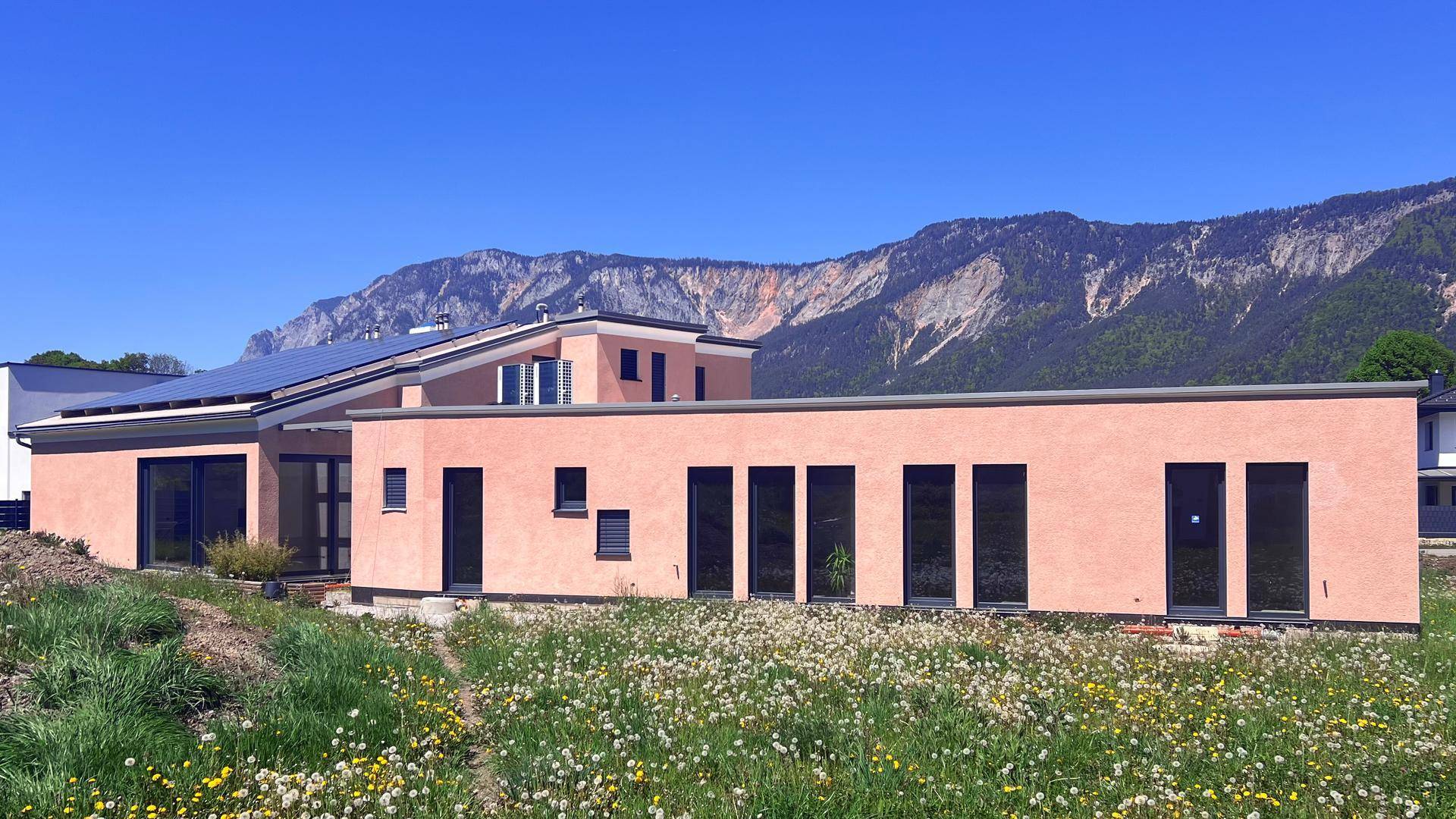 Modernes Haus mit rosafarbener Fassade, Flachdach und Photovoltaikanlage vor grüner Berglandschaft.