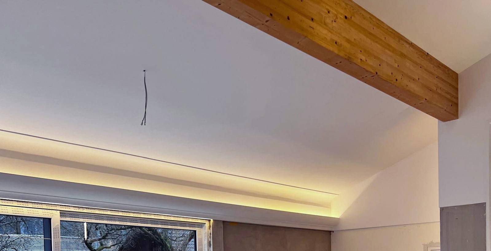 Decke mit sichtbarem Holzbalken und indirekter Beleuchtung über einem großen Fenster.
