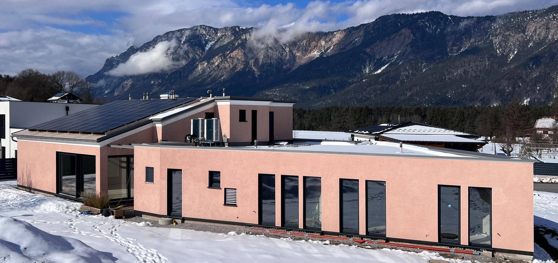Modernes Haus mit rosafarbener Fassade, Flachdach und Photovoltaikanlage vor verschneiter Berglandschaft.